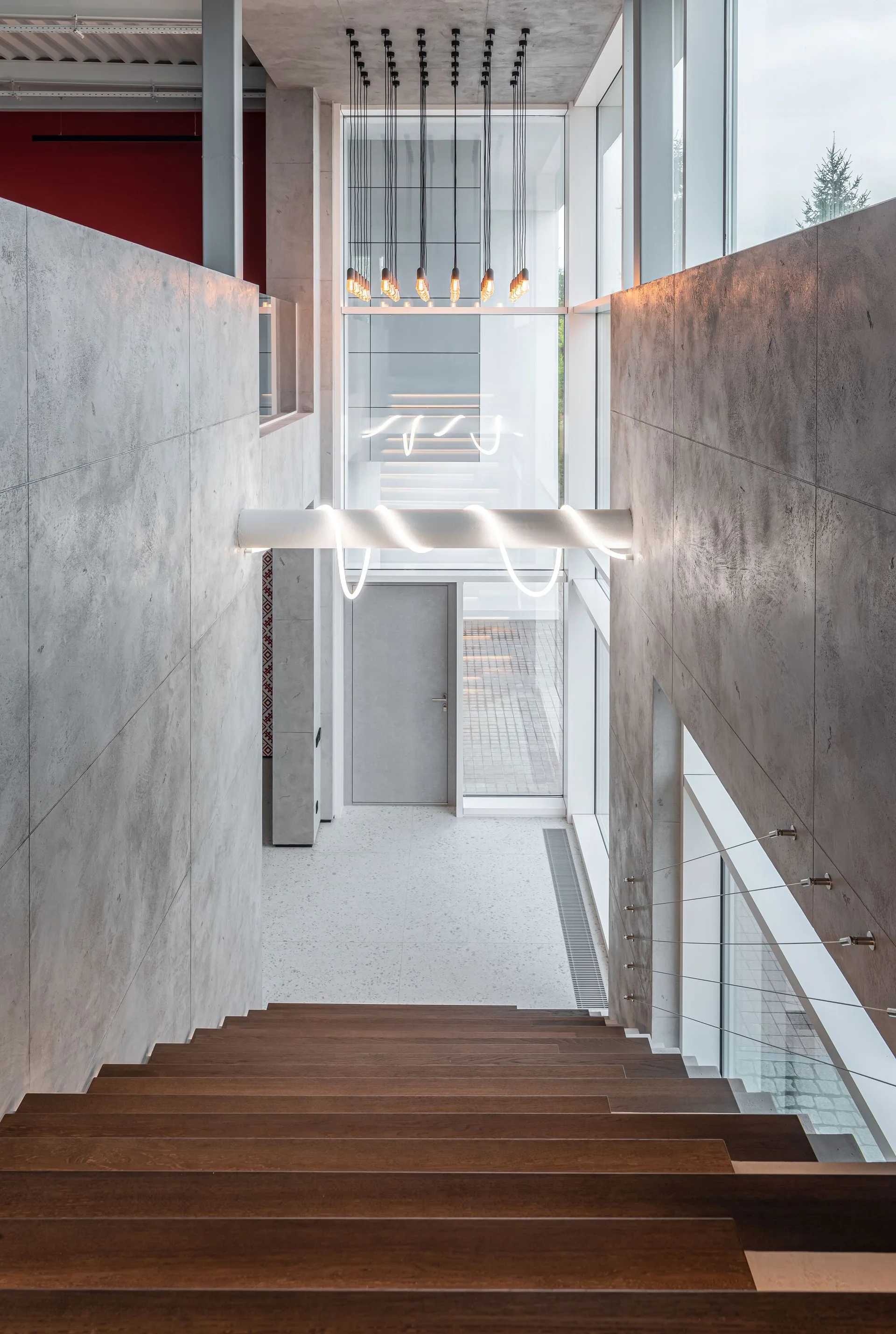 A view down wooden stairs flanked by concrete panels leading to a landing with large windows, hanging light bulbs, and a minimalist gray door.
