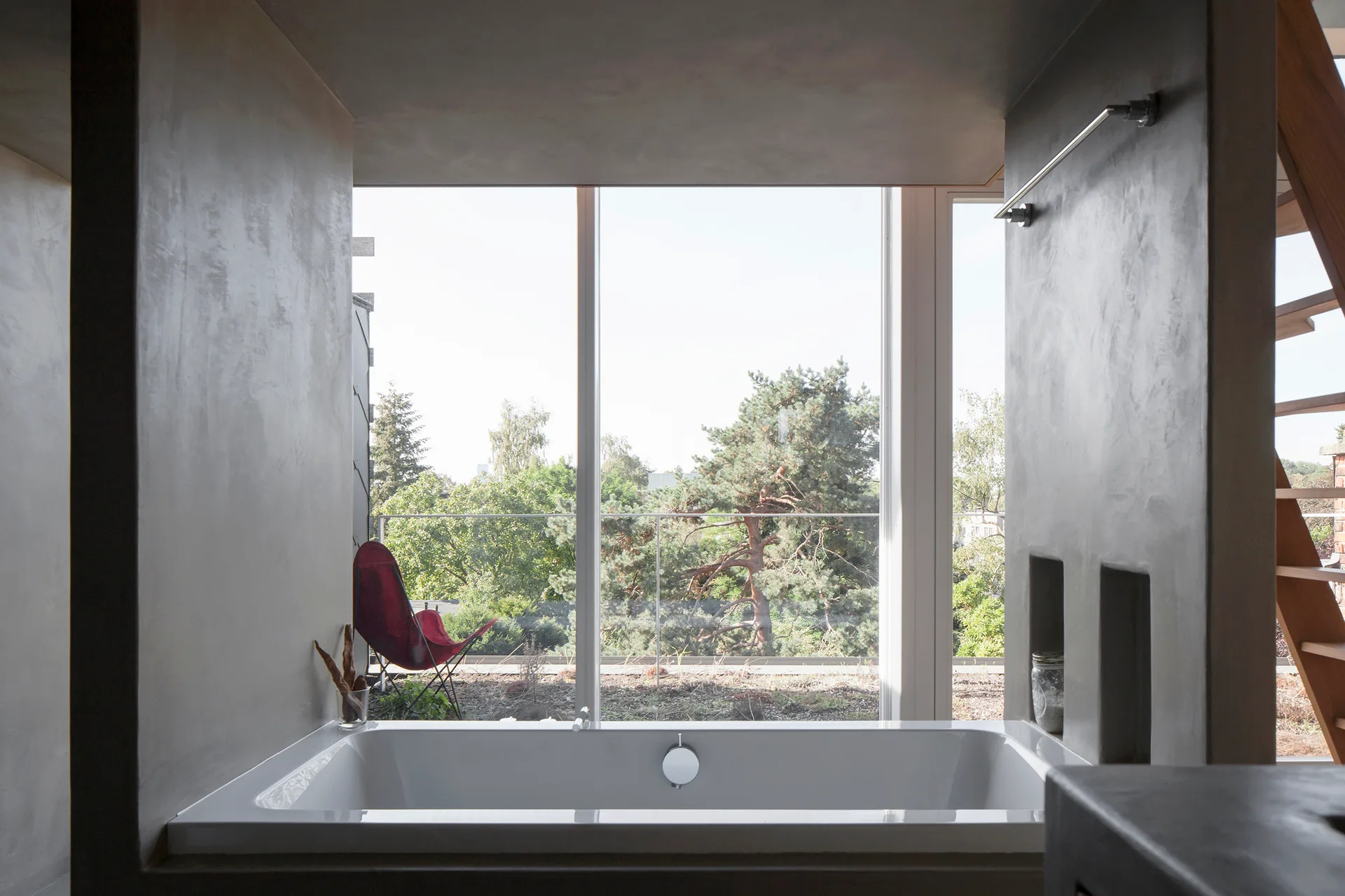 A white rectangular bathtub is set between smooth gray concrete-finish walls with a large glass window looking out onto a green terrace.