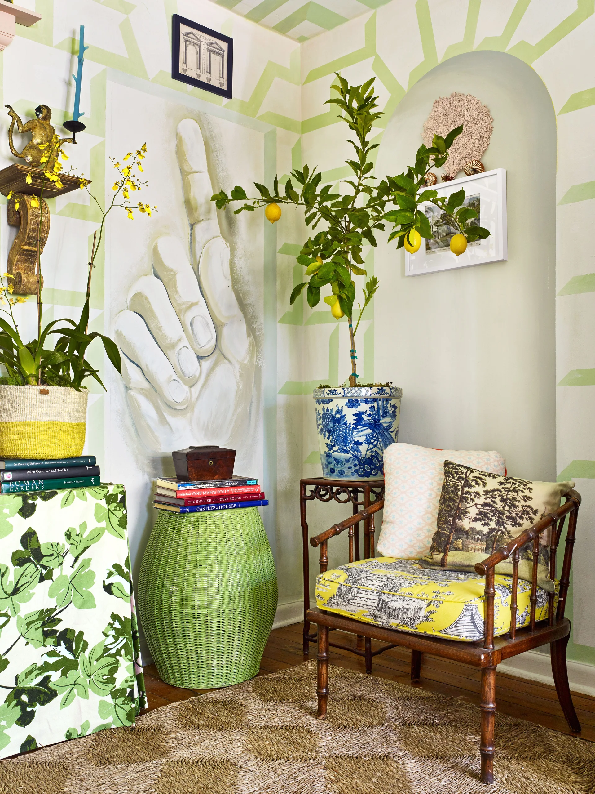 An eclectic corner features a wooden armchair with yellow patterned cushions, a potted lemon tree, a green wicker stool, and a hand mural.
