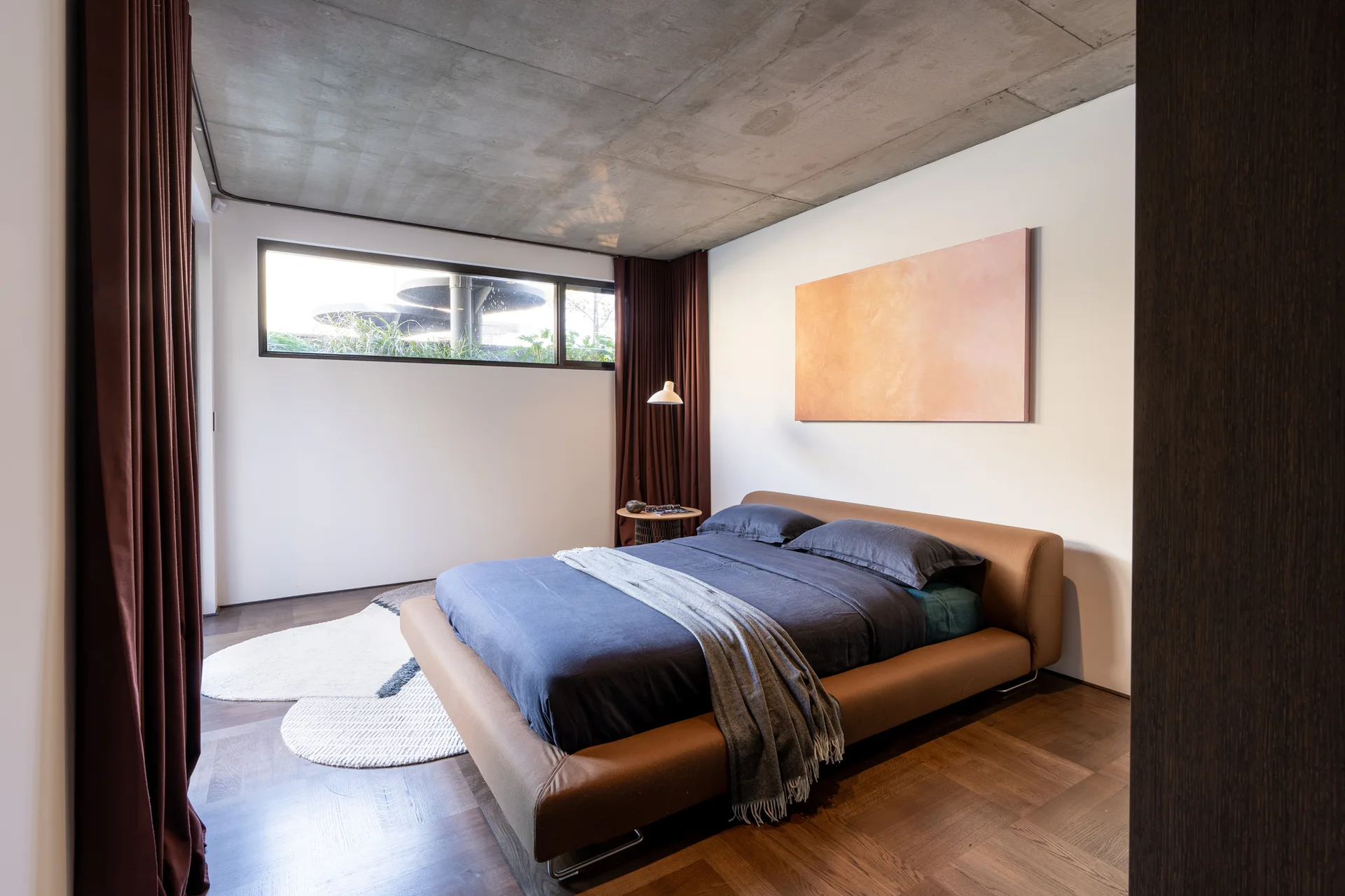 A modern bedroom features a low-profile brown upholstered bed, dark bedding, an exposed concrete ceiling, and long deep-red drapes over parquet flooring.