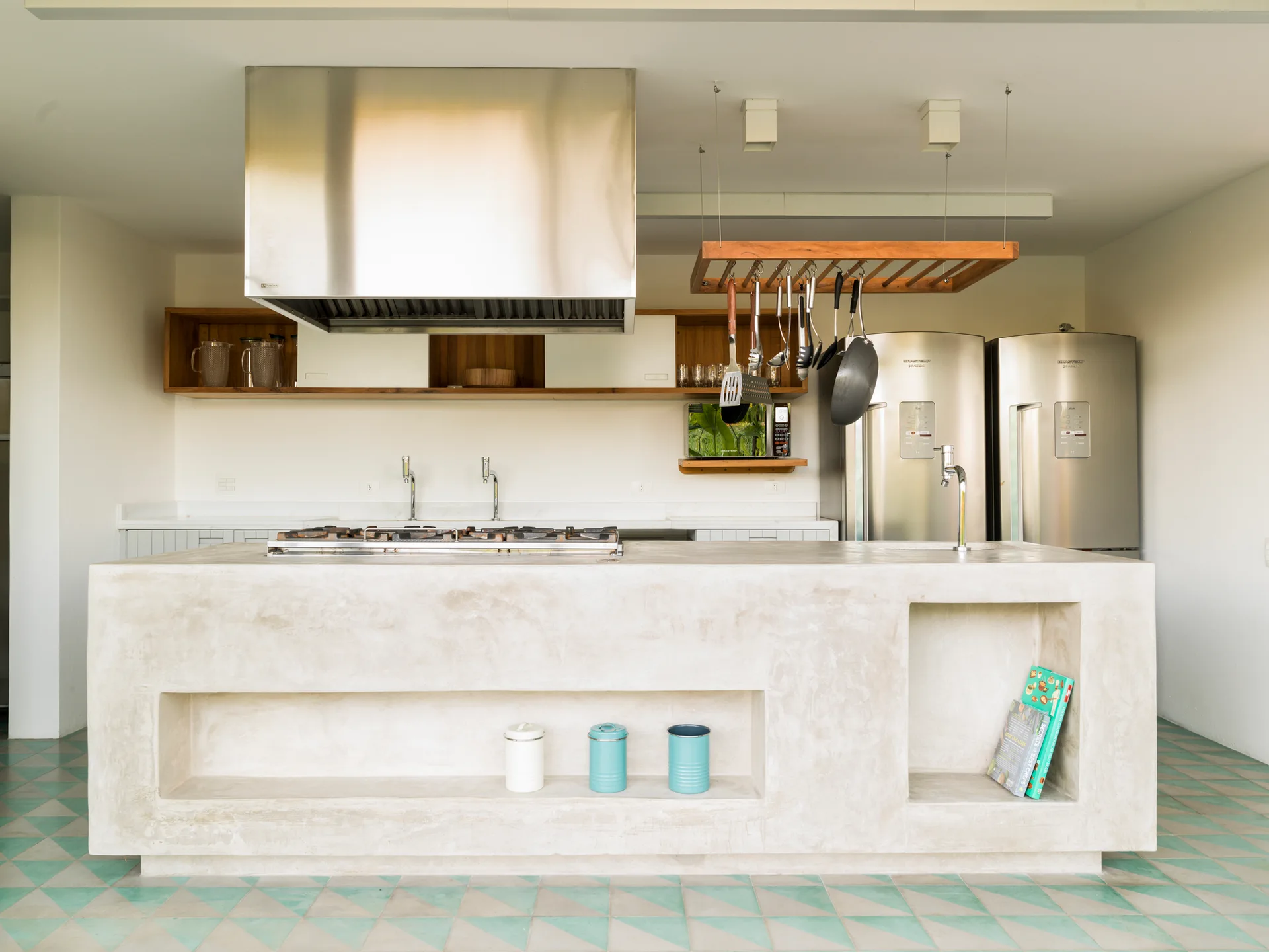 A modern kitchen featuring a large concrete island with integrated storage niches, a stainless steel range hood, and teal geometric floor tiles.