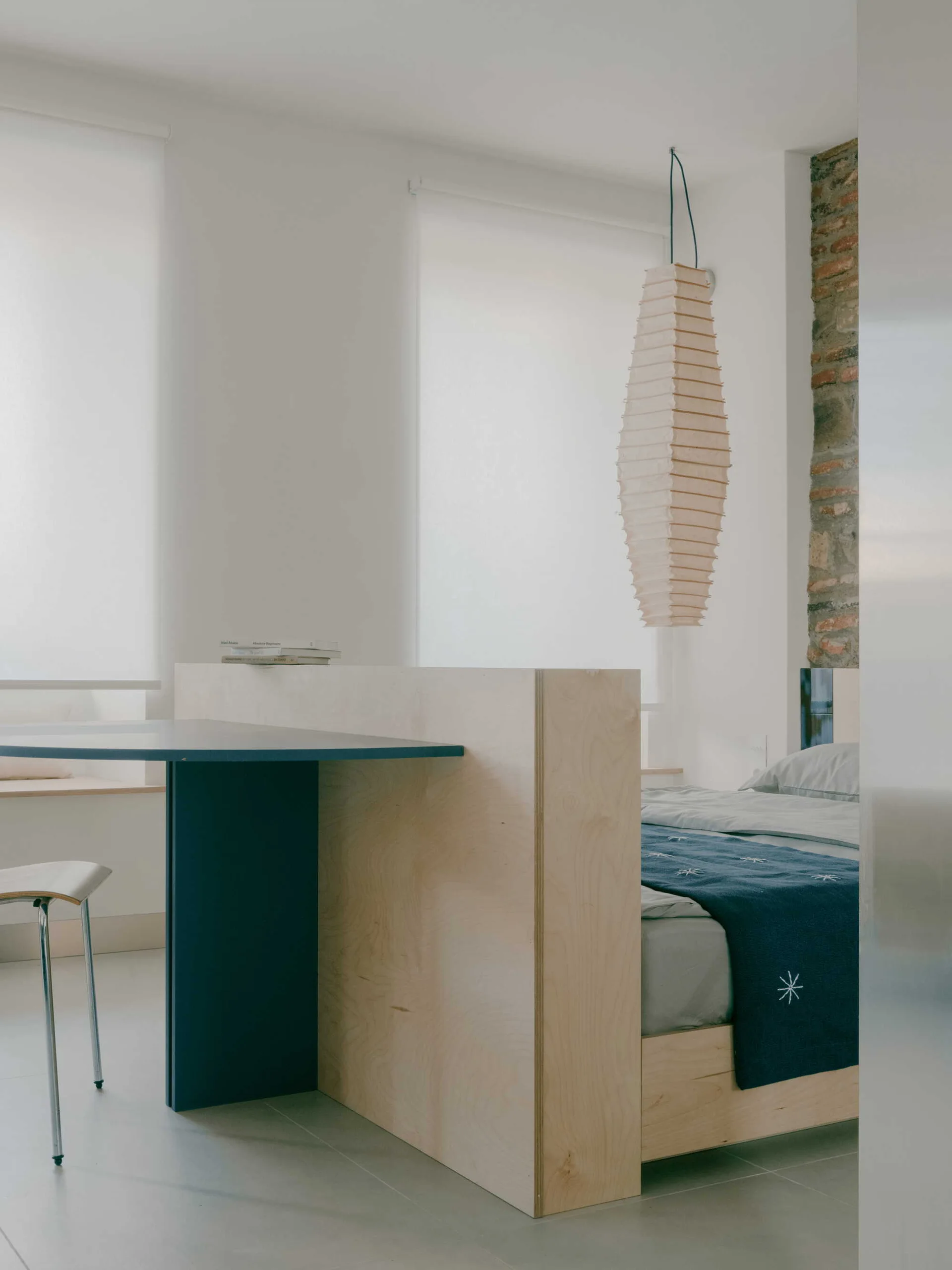 A minimalist bedroom features a custom plywood bed frame with an integrated blue desk, accompanied by a tall paper pendant light and exposed brick.