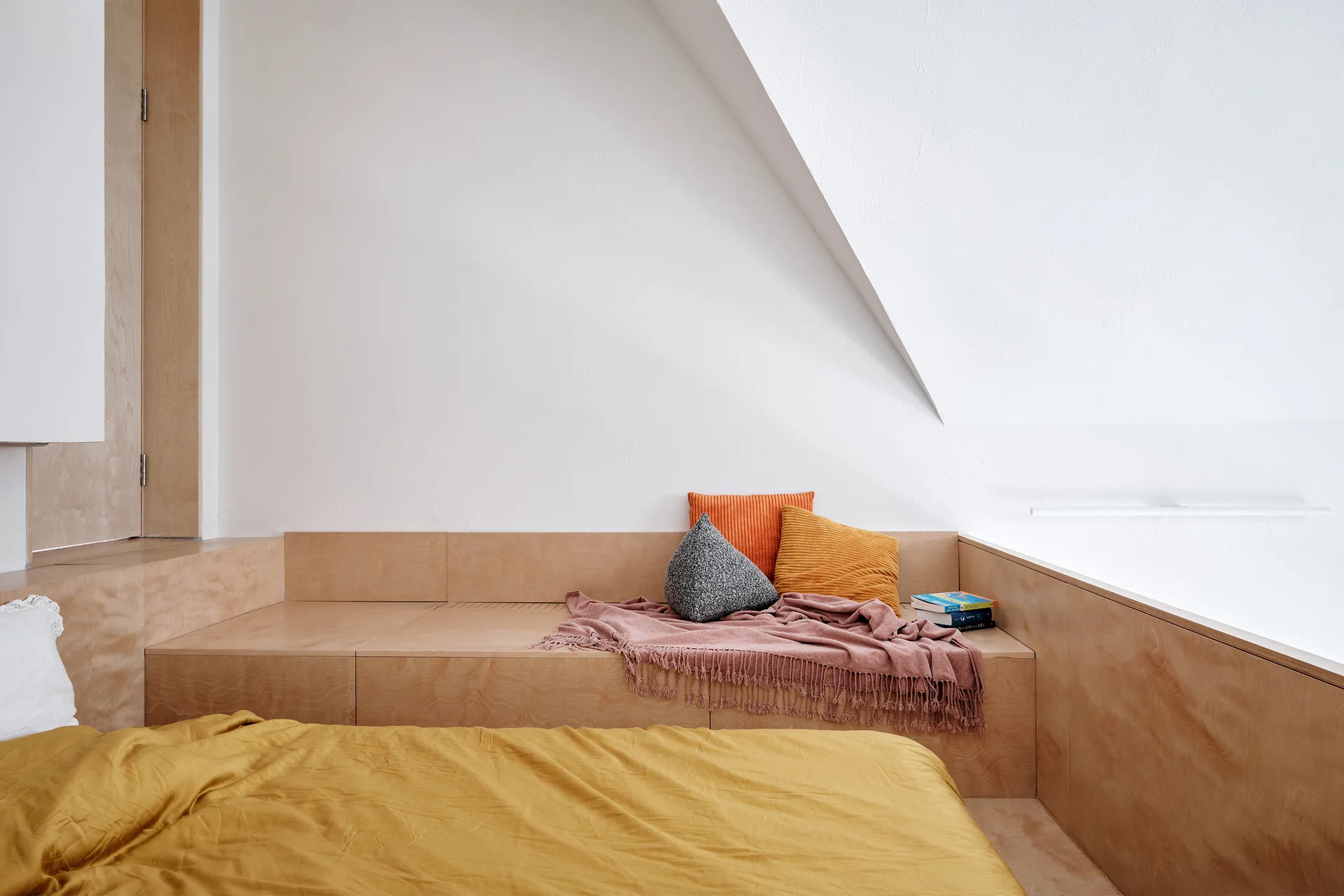 A minimalist attic bedroom features a built-in light wood platform bed with ochre yellow linens and colorful throw pillows under a sloped white ceiling.