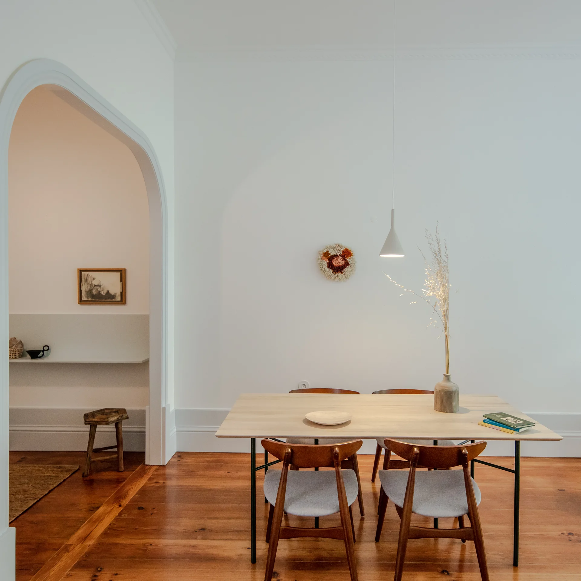 A minimalist dining room features a light wood table with mid-century chairs under a white pendant light next to an arched doorway.