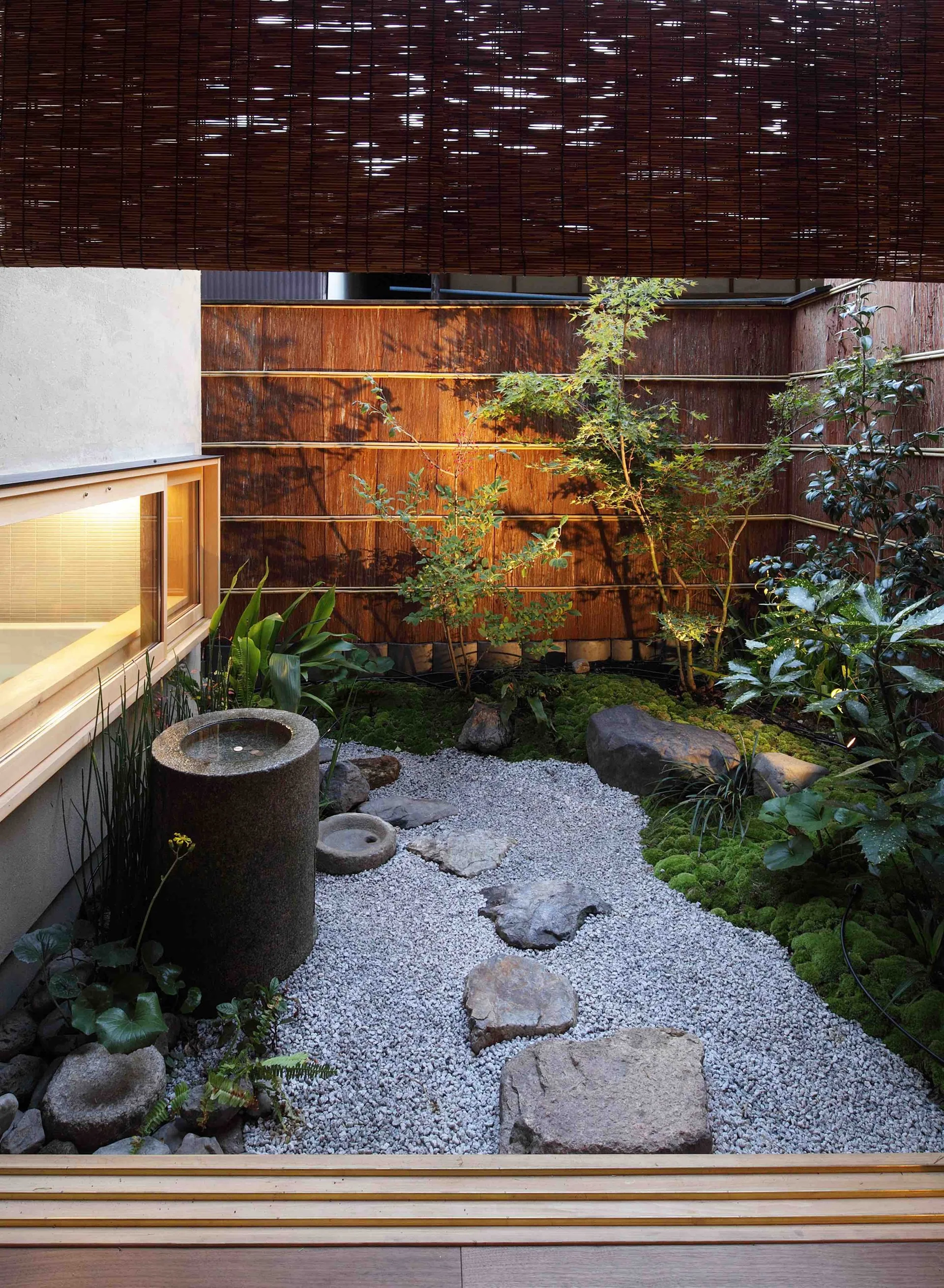 A small Japanese courtyard garden features a gravel path with stepping stones, green moss, a stone water basin, and a dark wood privacy fence.