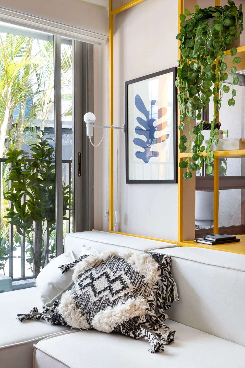 A white daybed features a textured black and white throw pillow next to a yellow metal shelving unit displaying plants and framed art.