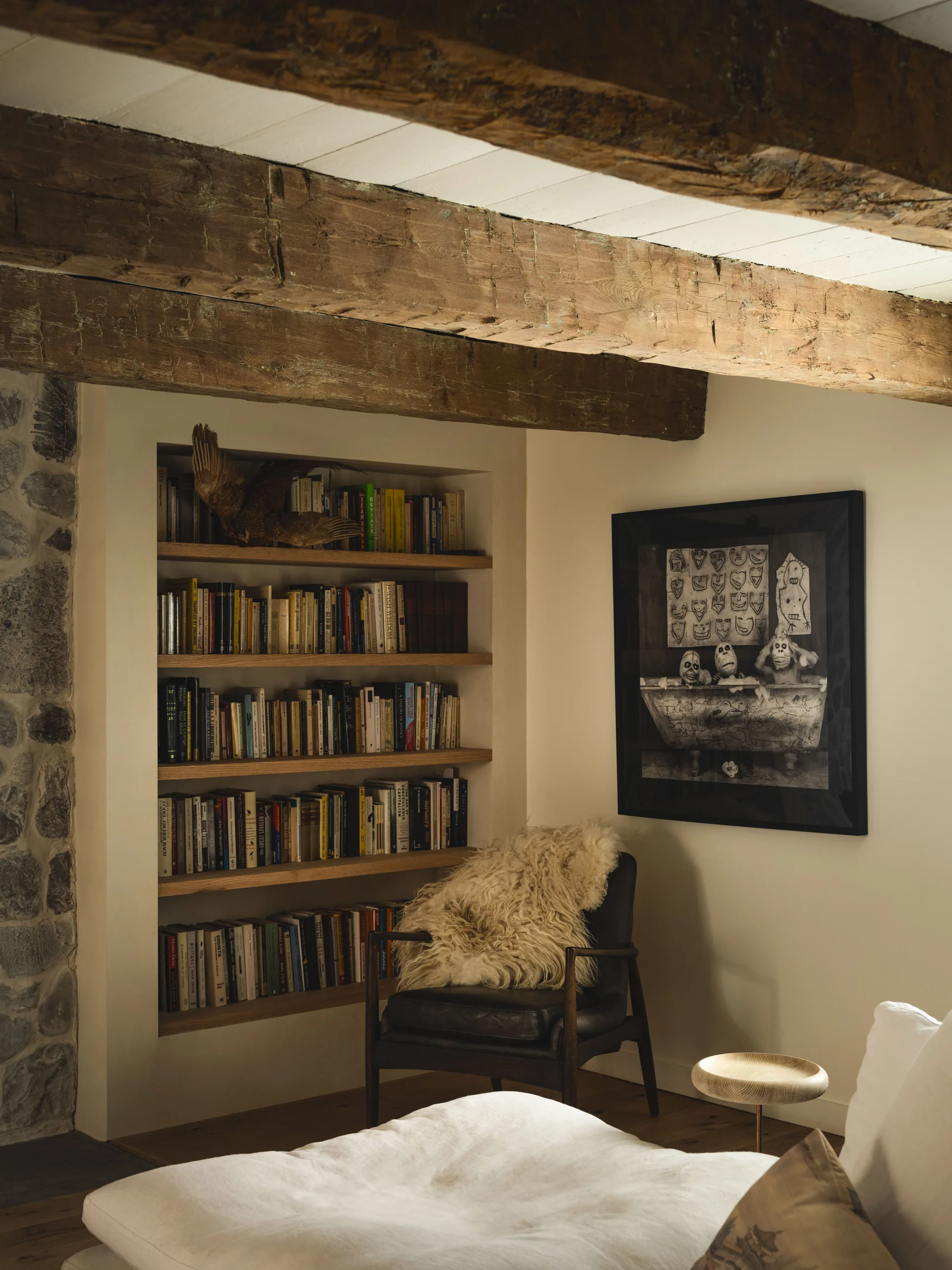 A cozy rustic reading corner with a built-in bookshelf, a dark leather armchair covered in a fluffy sheepskin, and heavy wood ceiling beams.