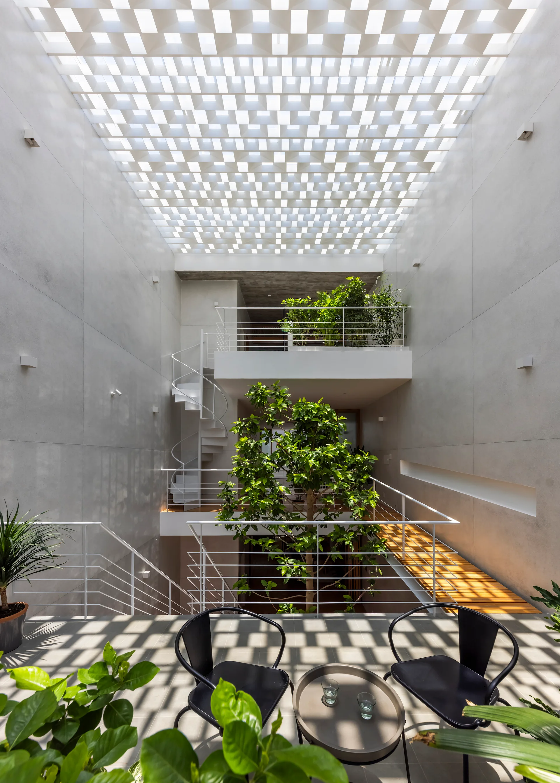 A multi-level modern atrium featuring a central green tree, a white spiral staircase, and a lattice ceiling casting grid shadows.