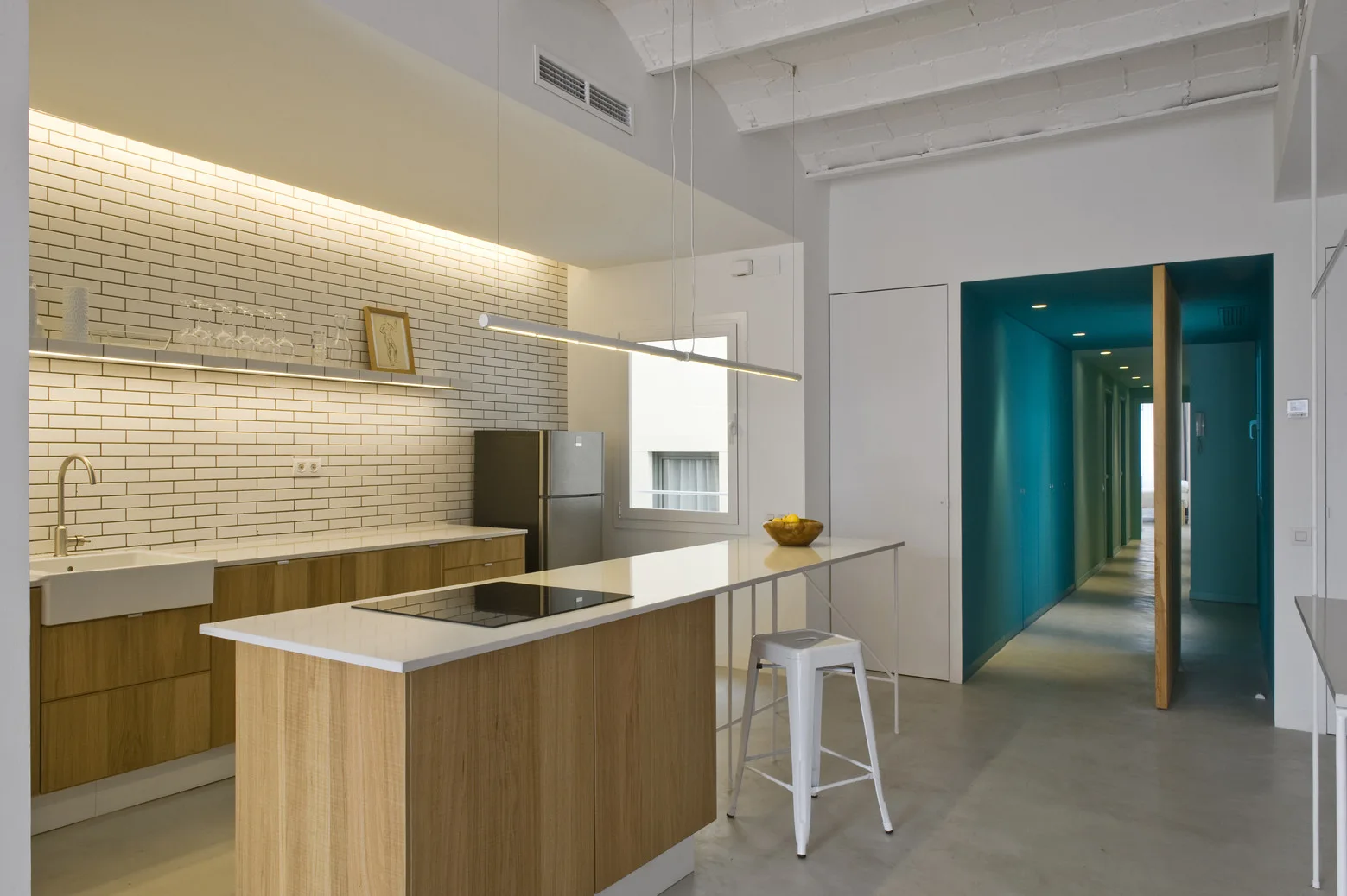 A modern kitchen featuring a wood-toned island with a white countertop, white subway tile, and a view into a deep blue hallway.