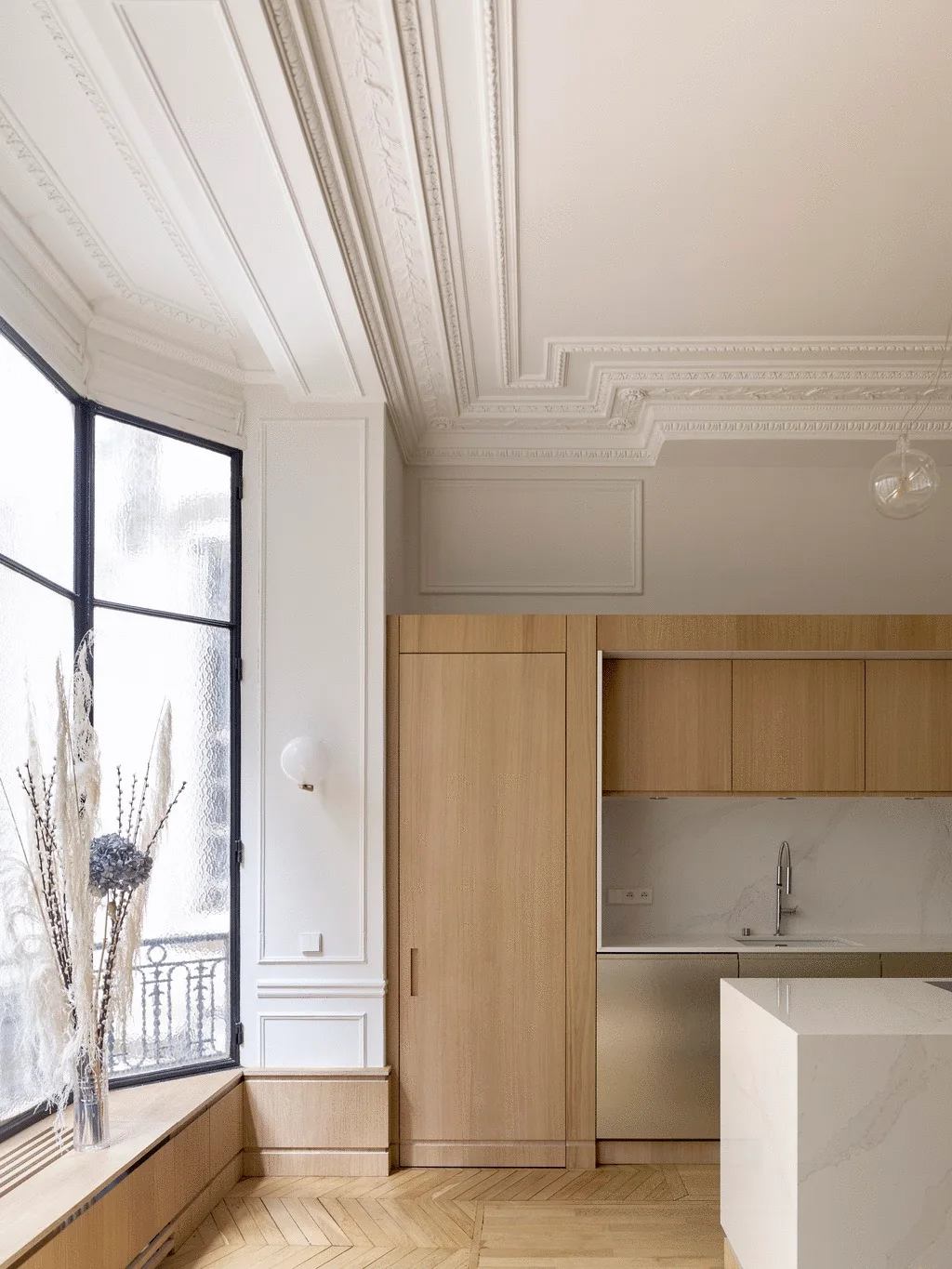 A modern kitchen with light wood cabinetry and a marble backsplash sits beneath a highly detailed white plaster ceiling near a bay window bench.