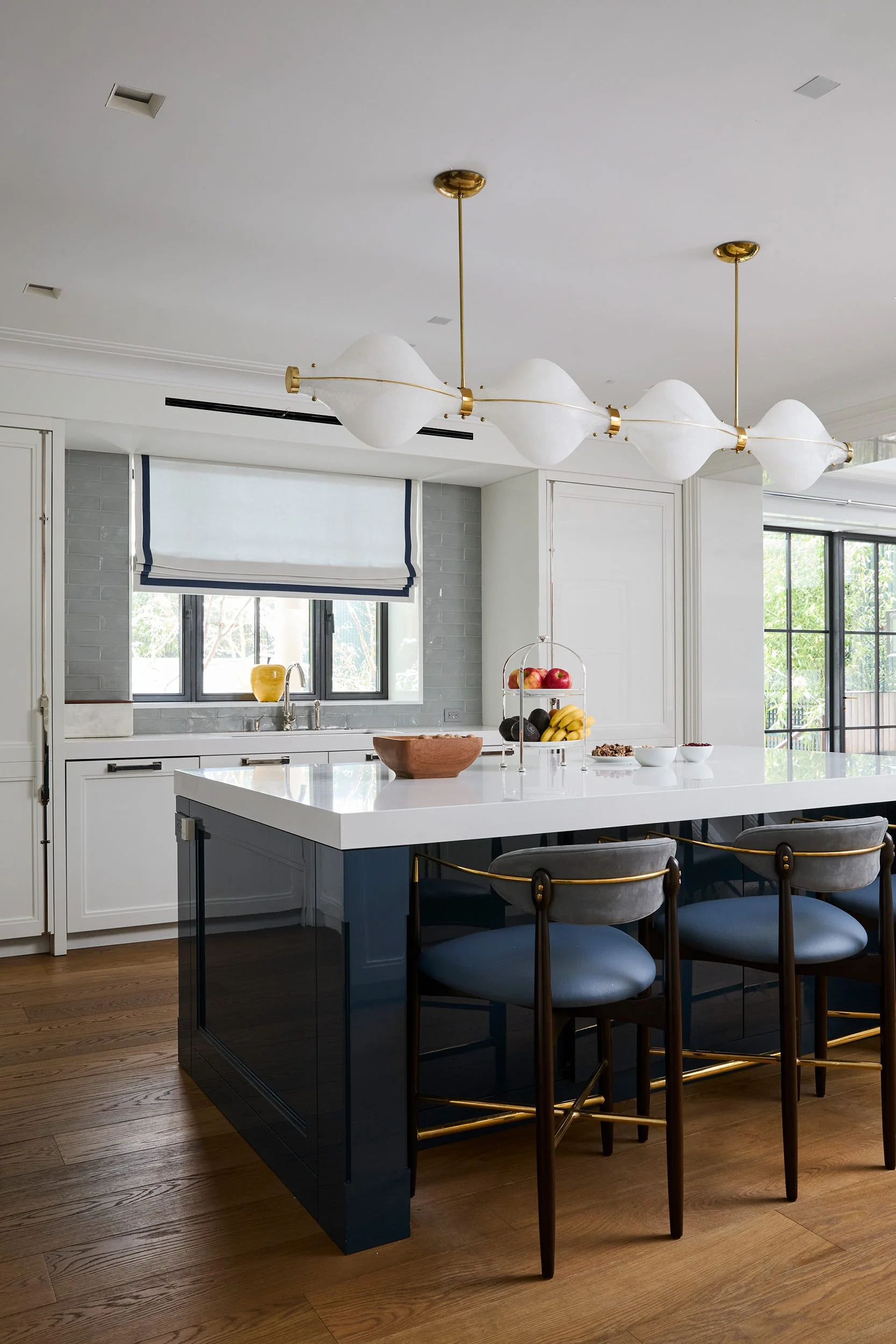 A contemporary kitchen features a glossy deep blue island with a white countertop, upholstered barstools, and a horizontal brass pendant light overhead.