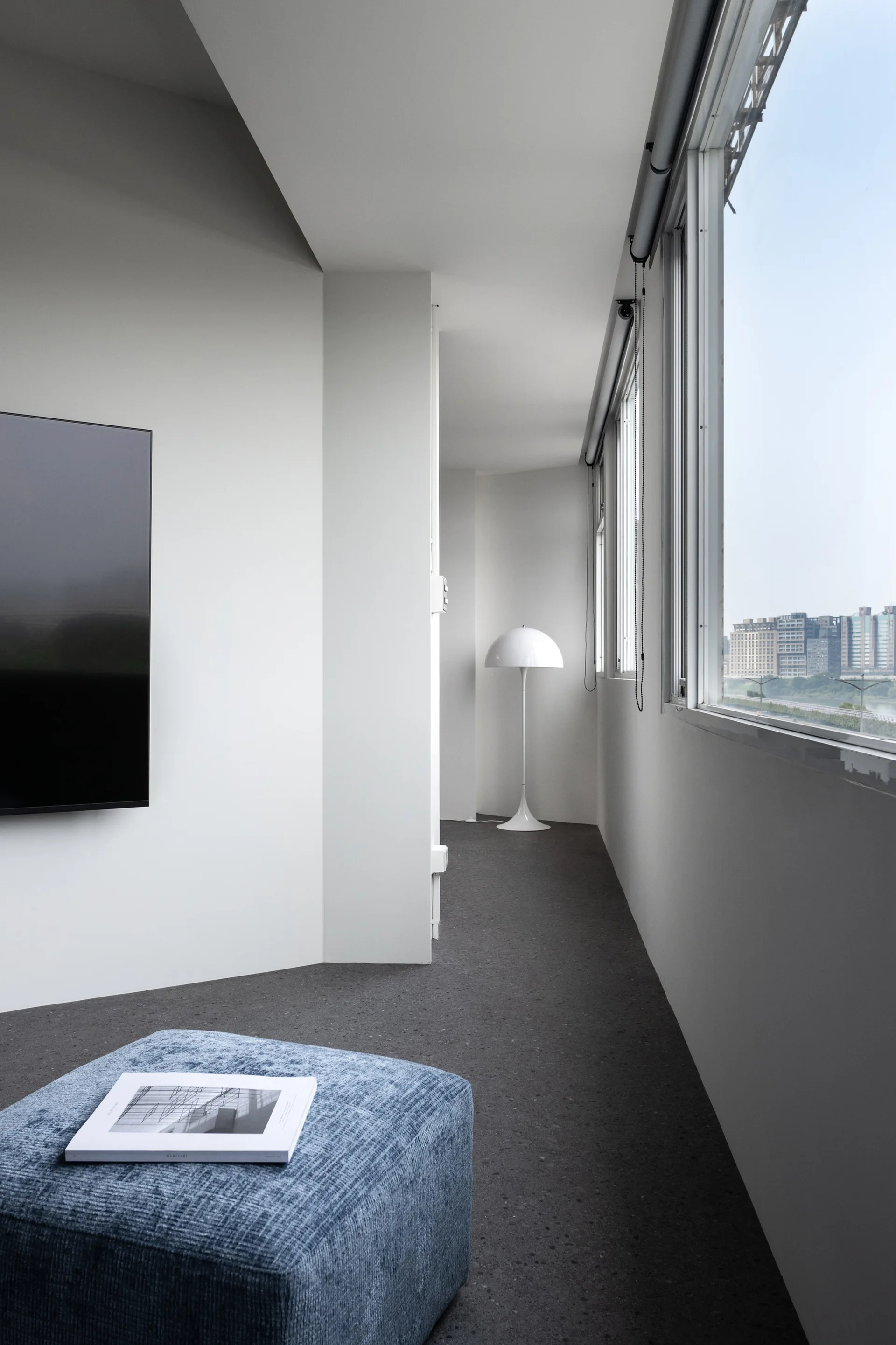 A minimalist white room features a textured blue ottoman in the foreground, a wall-mounted television, and a white dome floor lamp near a window.