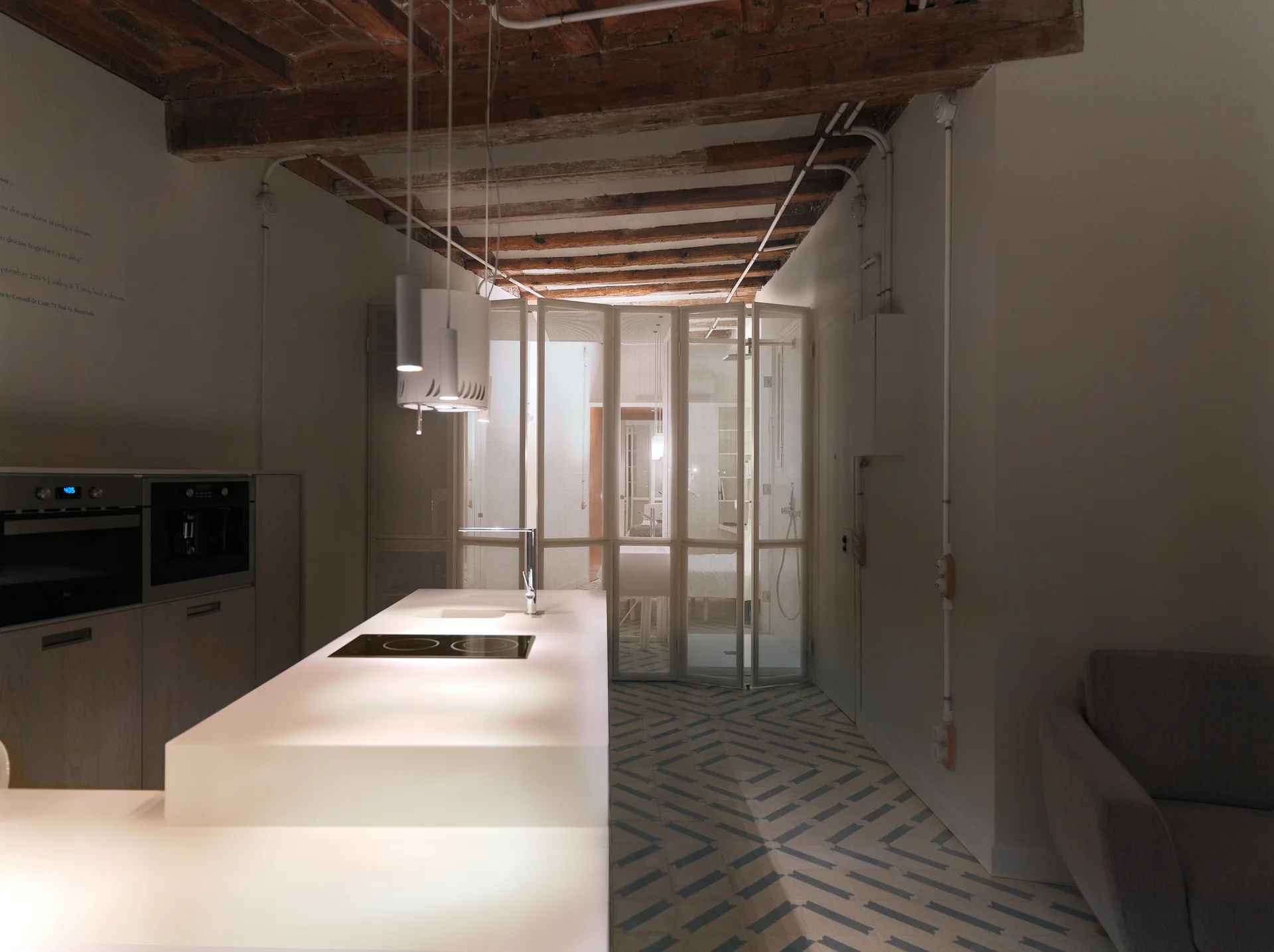 A modern kitchen featuring a glowing white island, exposed wood ceiling beams, patterned floor tiles, and a folding metal mesh screen partition.