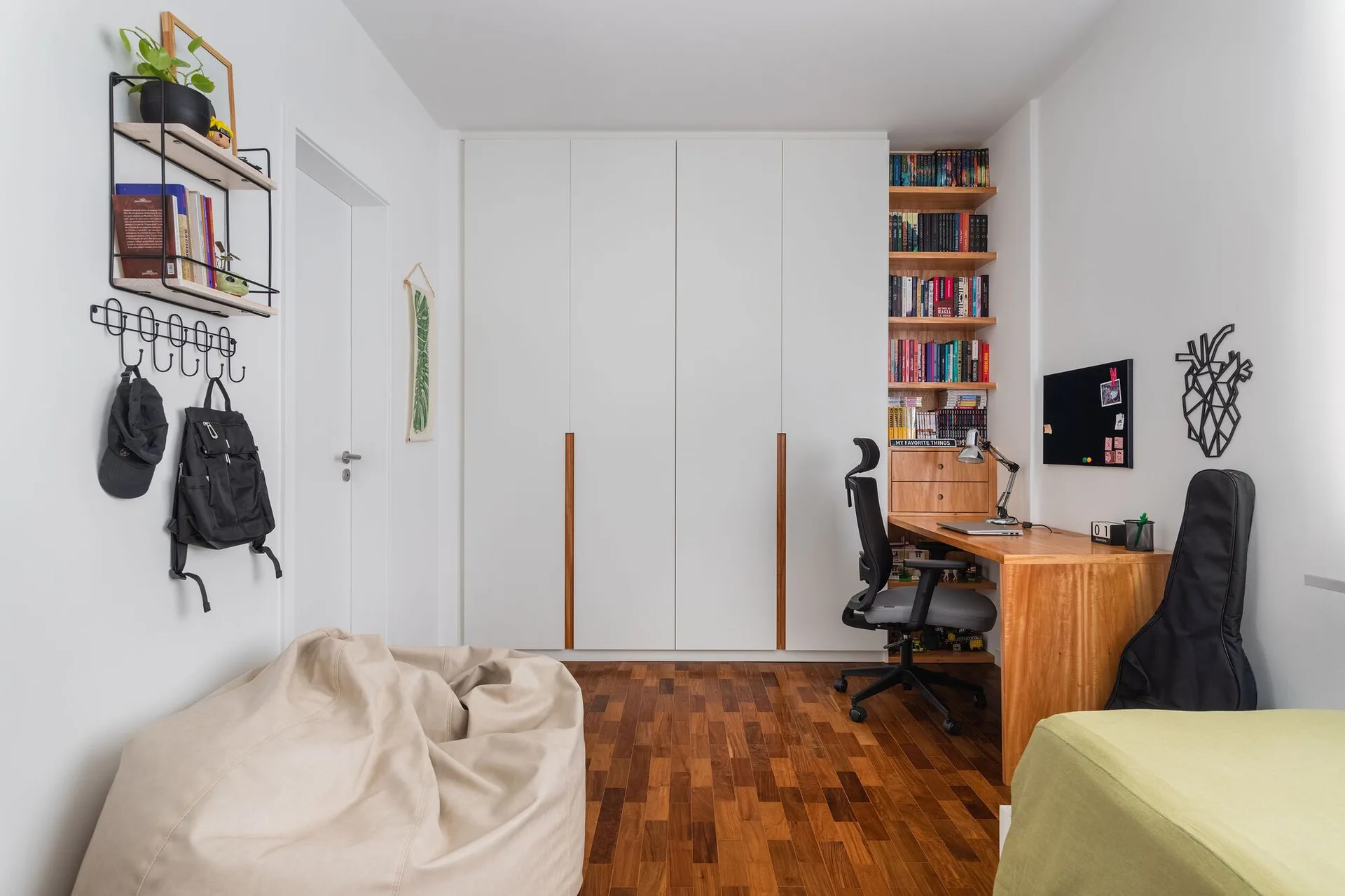 A white bedroom with parquet floors features a large built-in wardrobe, an integrated wood desk with bookshelves, and a beige beanbag.