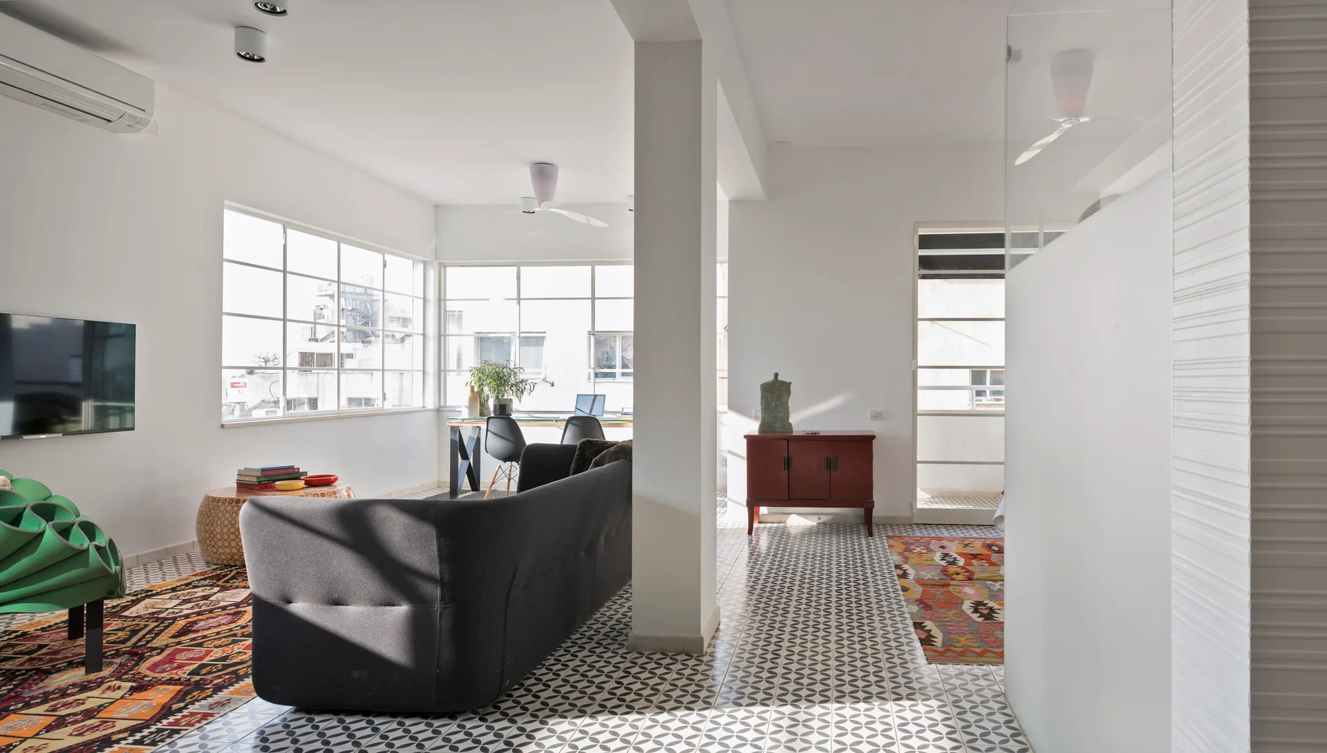 An open-plan living room featuring a dark sofa, geometric patterned floor tiles, large metal windows, and a white structural column.