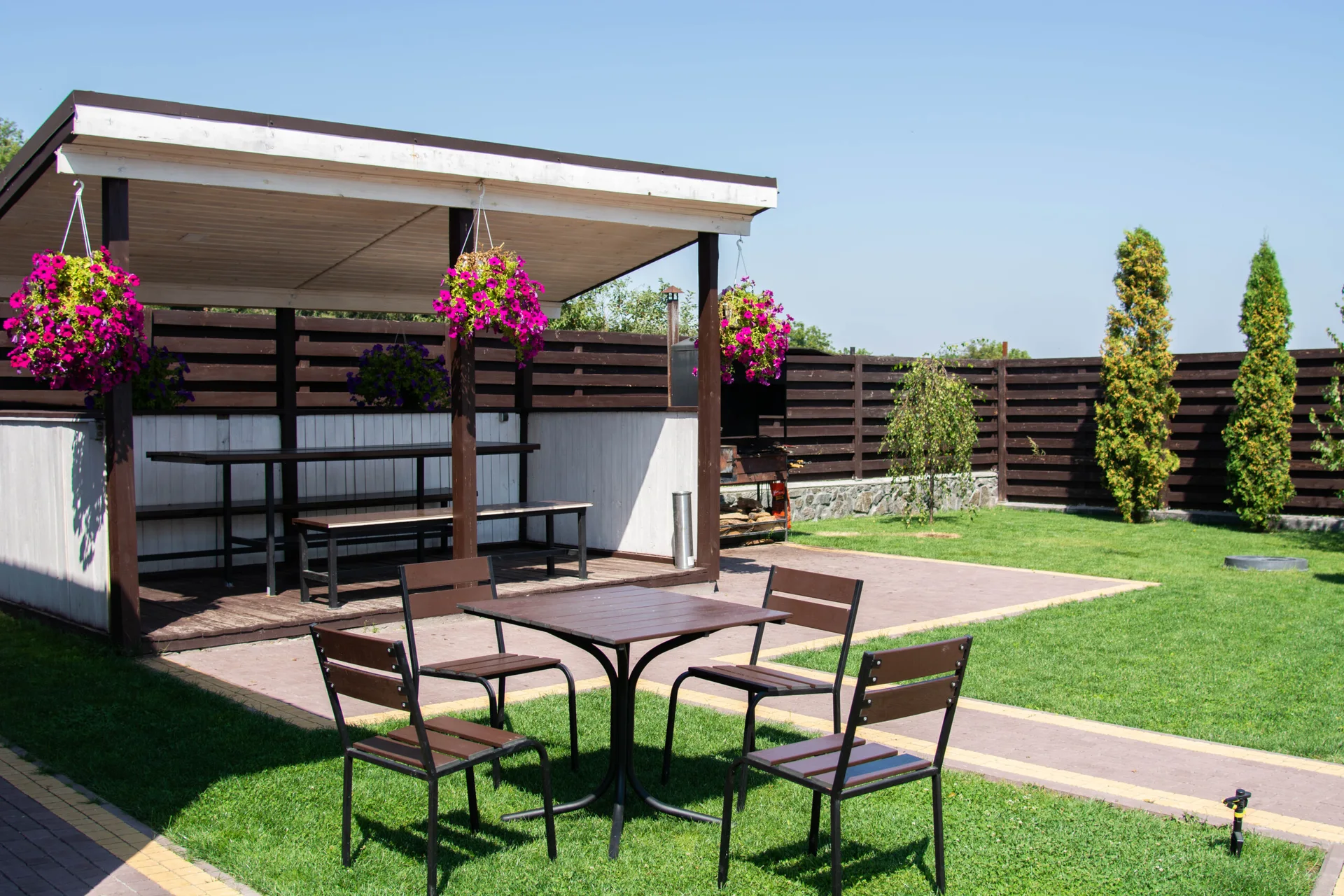 A backyard patio features a dark wood pergola with pink hanging flowers, a metal-framed dining set on a paved path, and a surrounding lawn with a fence.