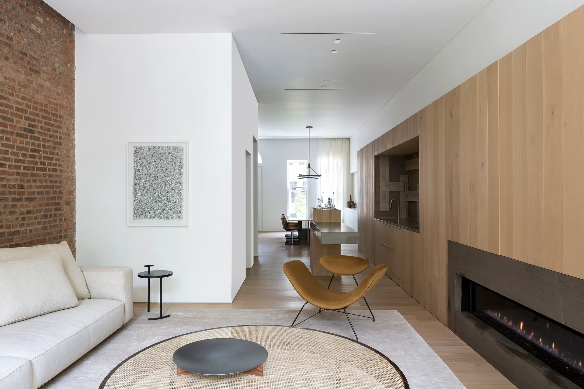 A minimalist living room featuring a light wood paneled wall with a fireplace, a cream sofa, and an exposed brick accent wall.