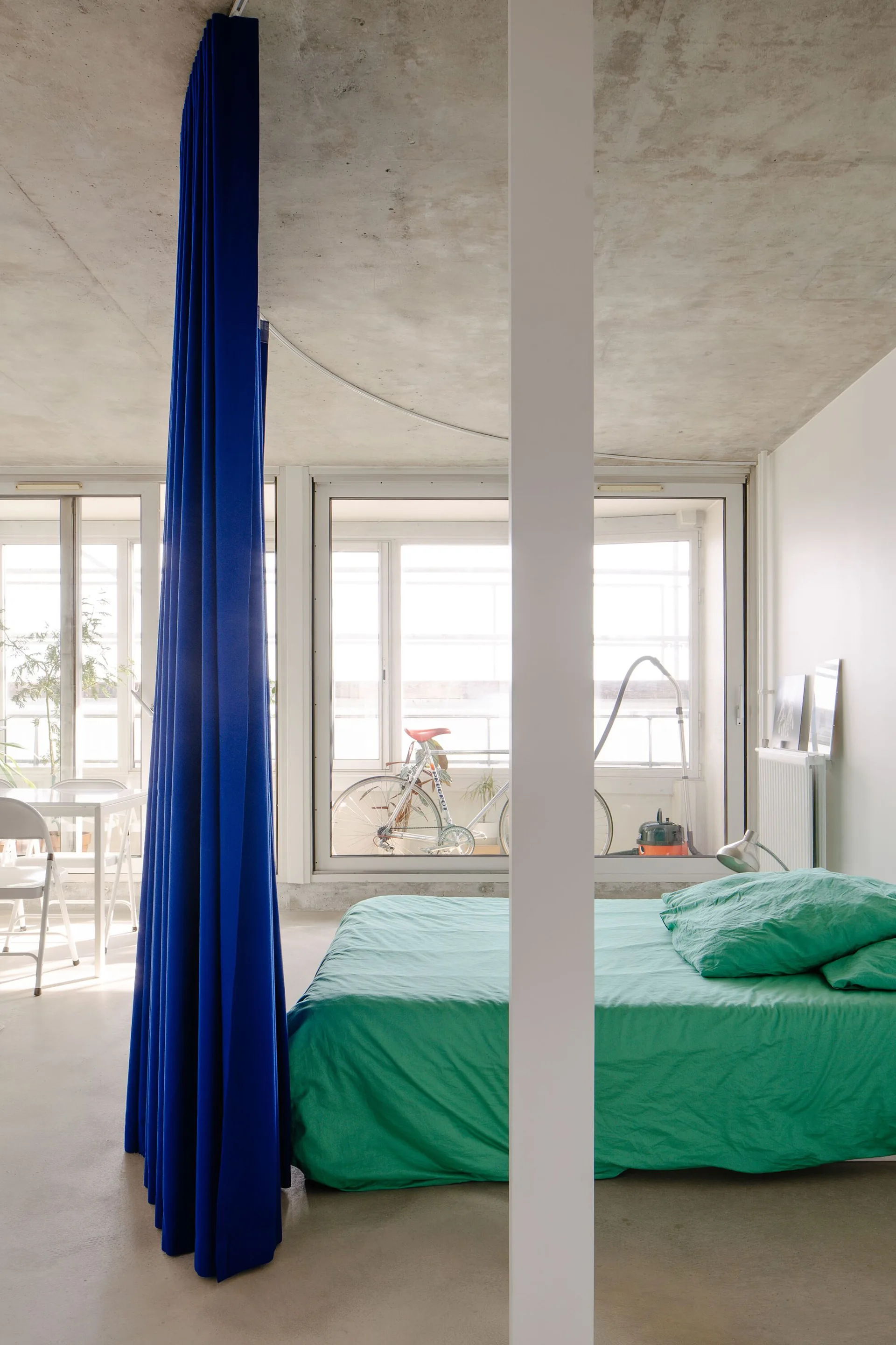 A minimalist studio bedroom features a green bed and a tall blue curtain partition under an exposed concrete ceiling with large windows.