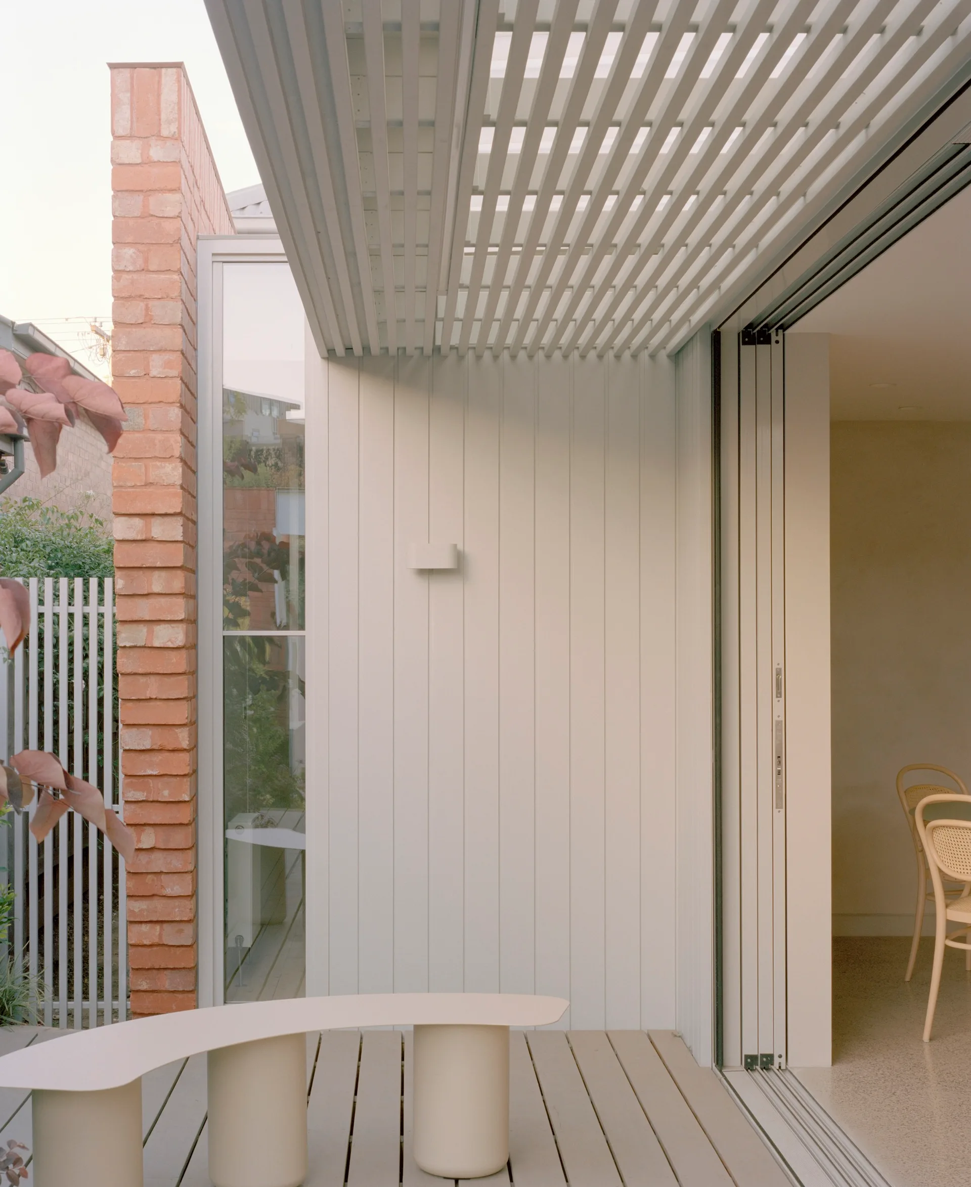 A minimalist outdoor patio featuring a curved beige bench, white vertical siding, a slatted overhead pergola, and an adjacent brick accent wall.