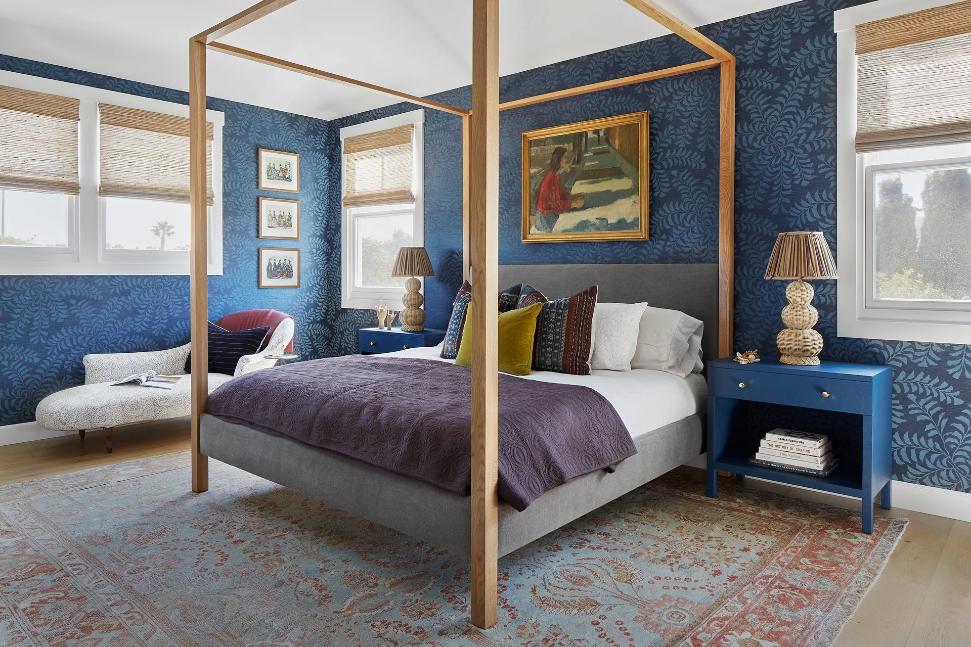 A bedroom featuring a wooden four-poster bed, blue leaf pattern wallpaper, matching blue nightstands, and woven roman shades.