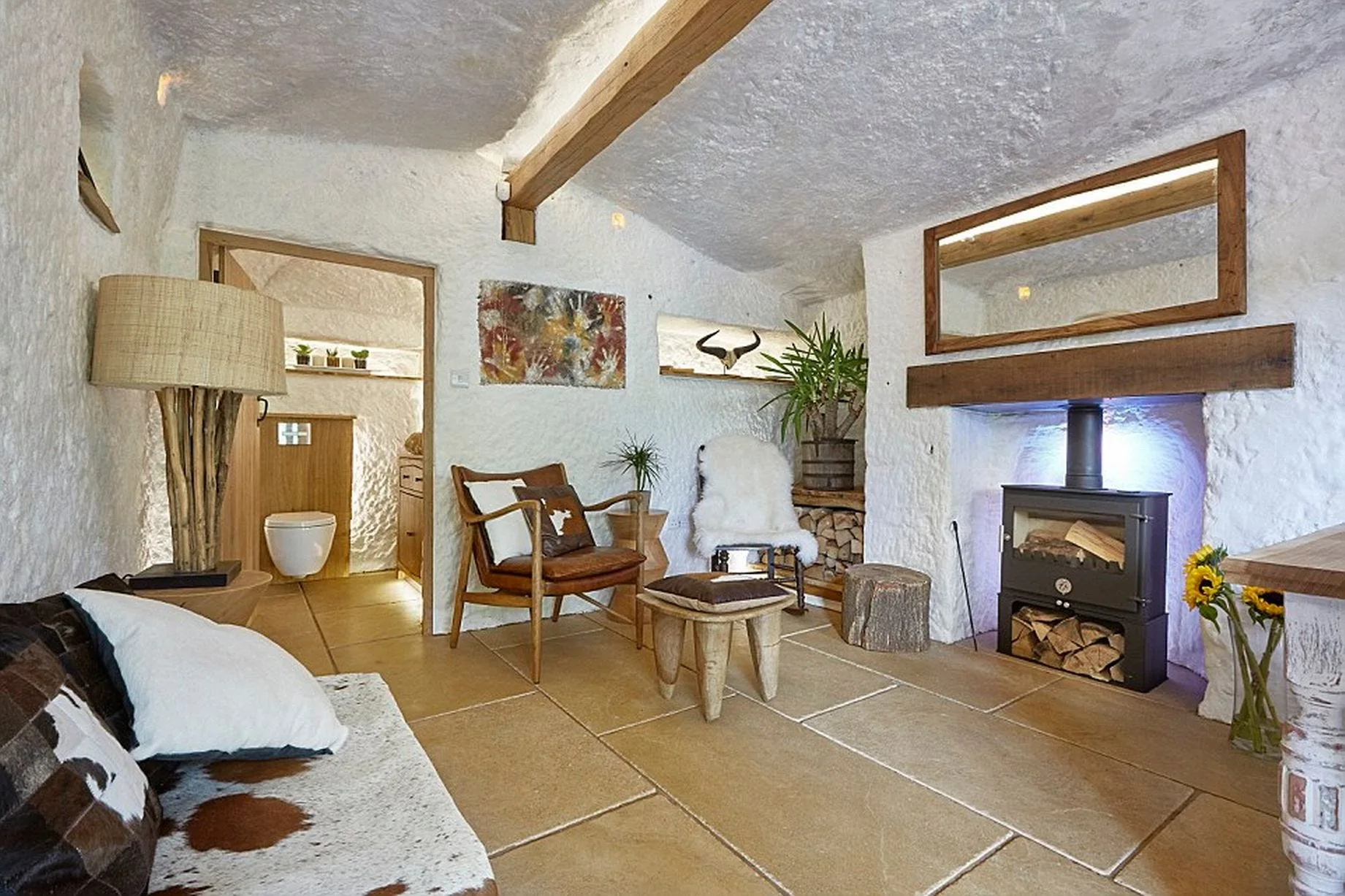 A rustic living room features white textured plaster walls, a black wood-burning stove, tan stone floor tiles, and a wooden ceiling beam.