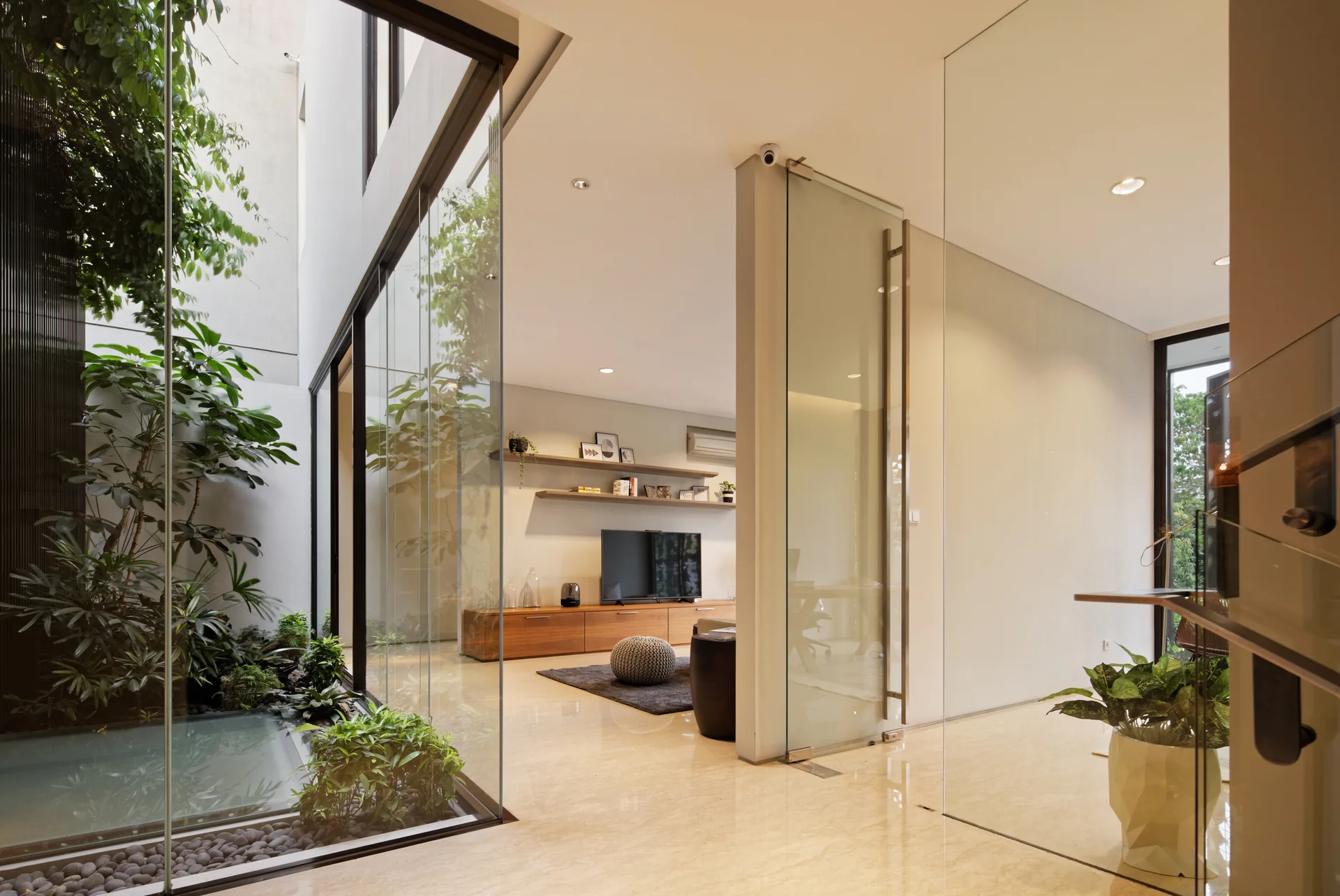 A modern living room with polished tile floors opens to a glass-enclosed indoor atrium featuring lush plants and a shallow water pool.