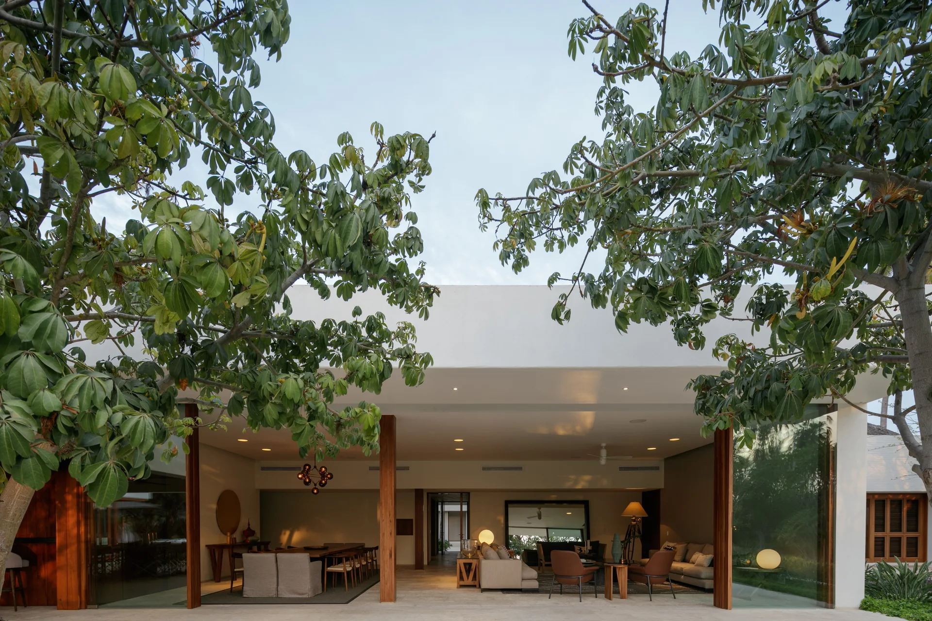 A wide covered patio features open-plan dining and living areas framed by large trees and supported by wooden pillars.