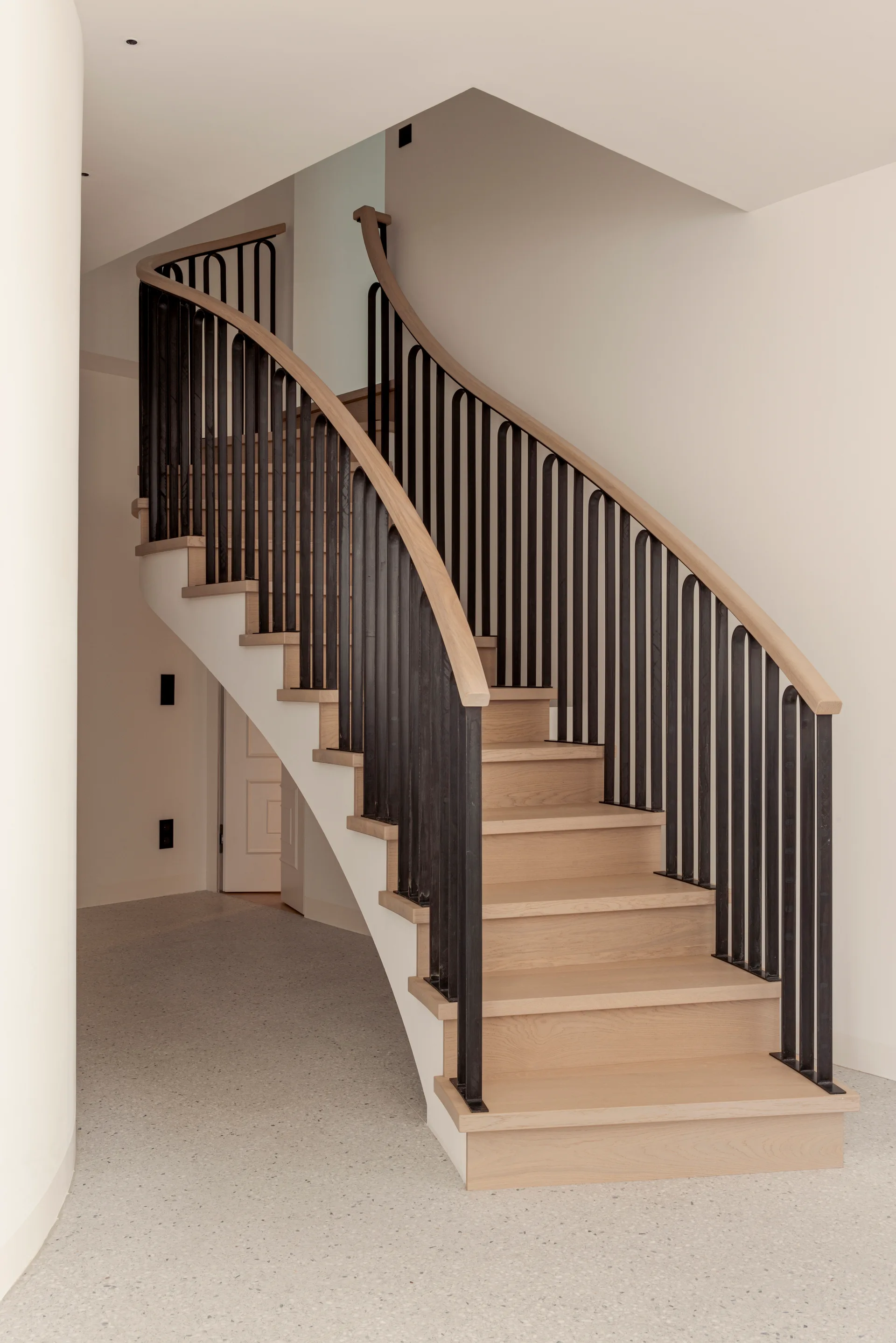 A curved staircase with light wood treads and a black metal spindle railing rises above a matte terrazzo floor.