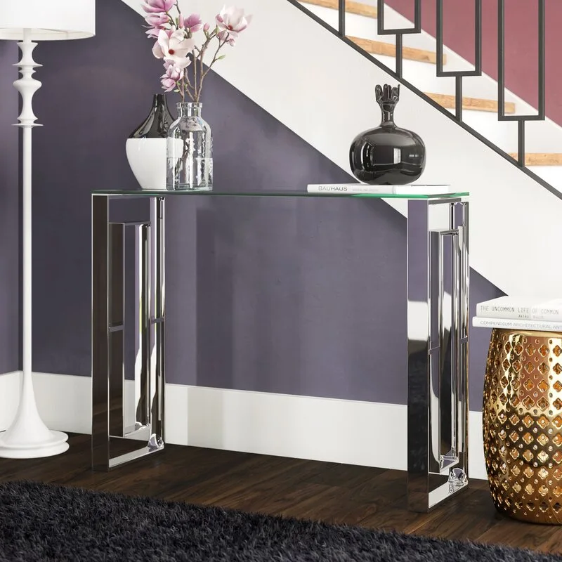 A glass and chrome console table rests against a deep purple wall beside a staircase, styled with vases, a white floor lamp, and a gold garden stool.