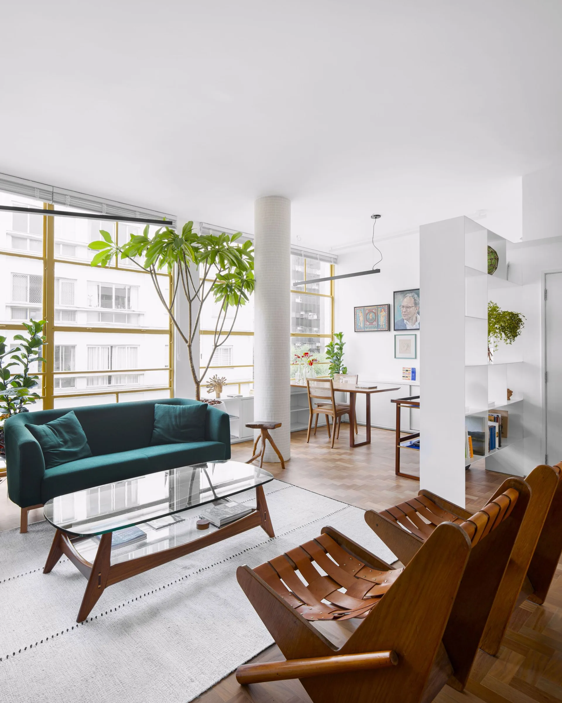 A mid-century modern living room features a dark green sofa, leather strap armchairs, a glass-topped coffee table, and a central tiled column.