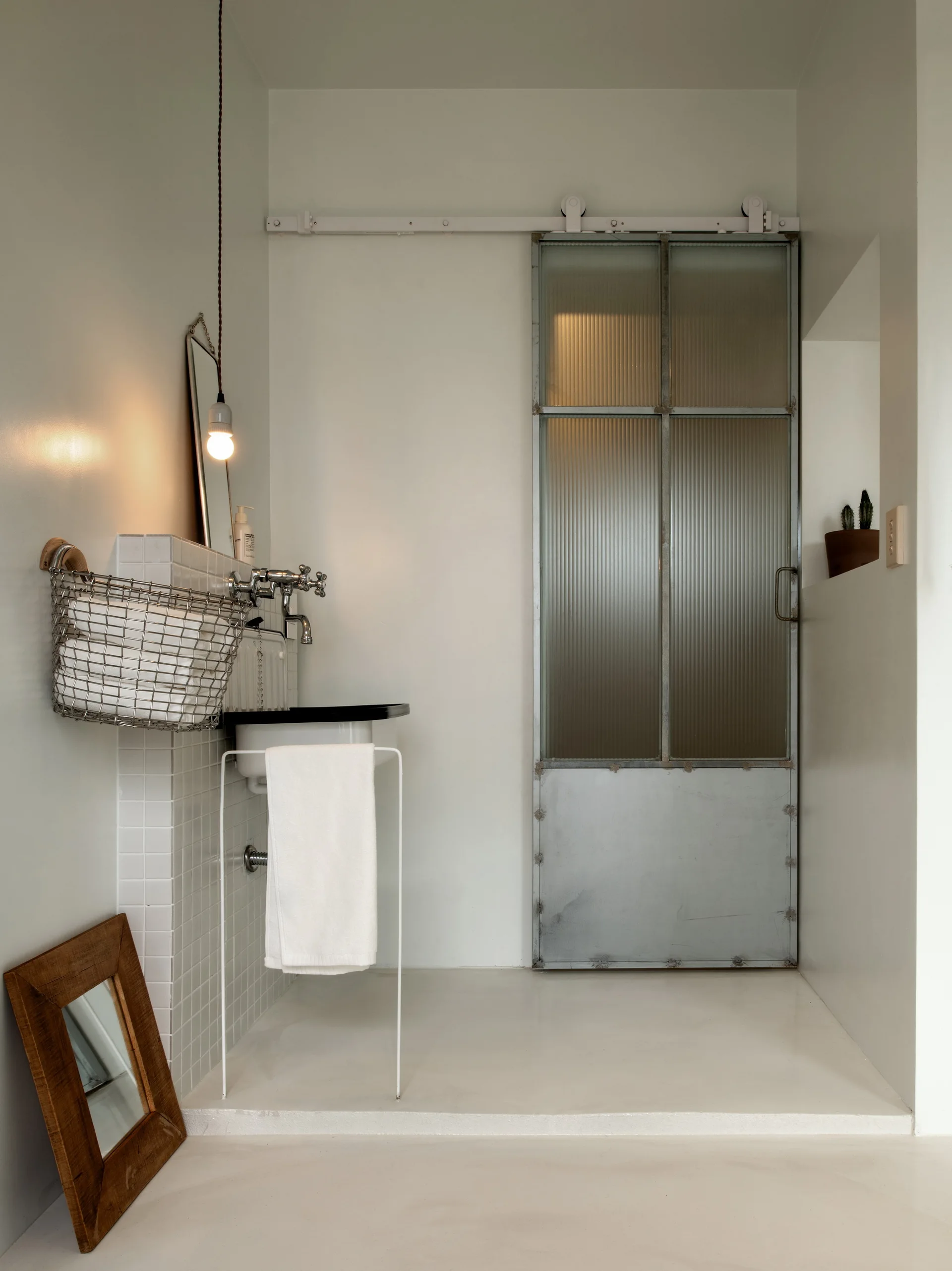 A minimalist white bathroom features a pedestal sink with small square tiles, a wire storage basket, and an industrial fluted glass sliding door.