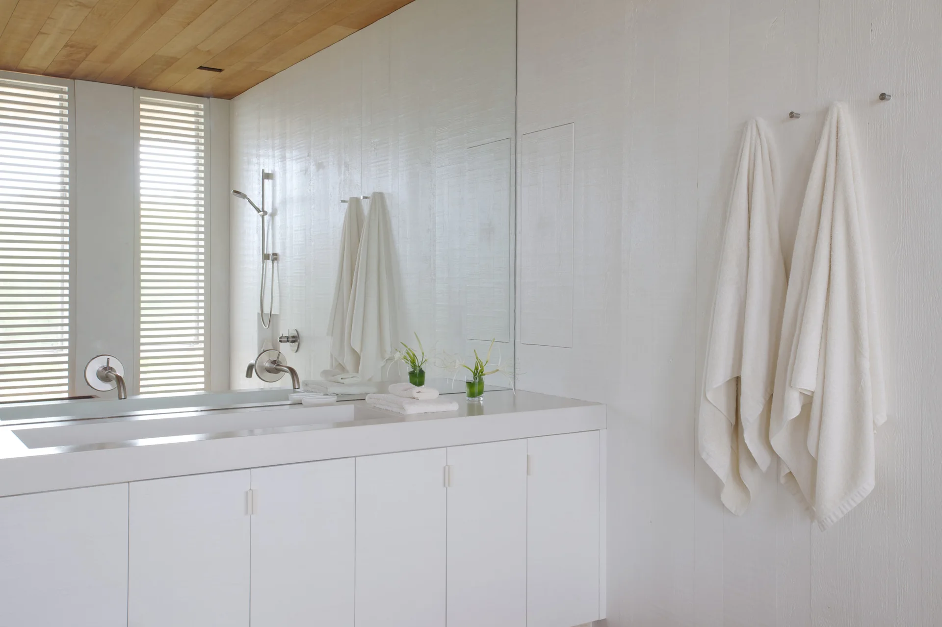 A minimalist white bathroom features an integrated vanity cabinet, vertical wood paneling, hanging white towels, and a natural wood plank ceiling.