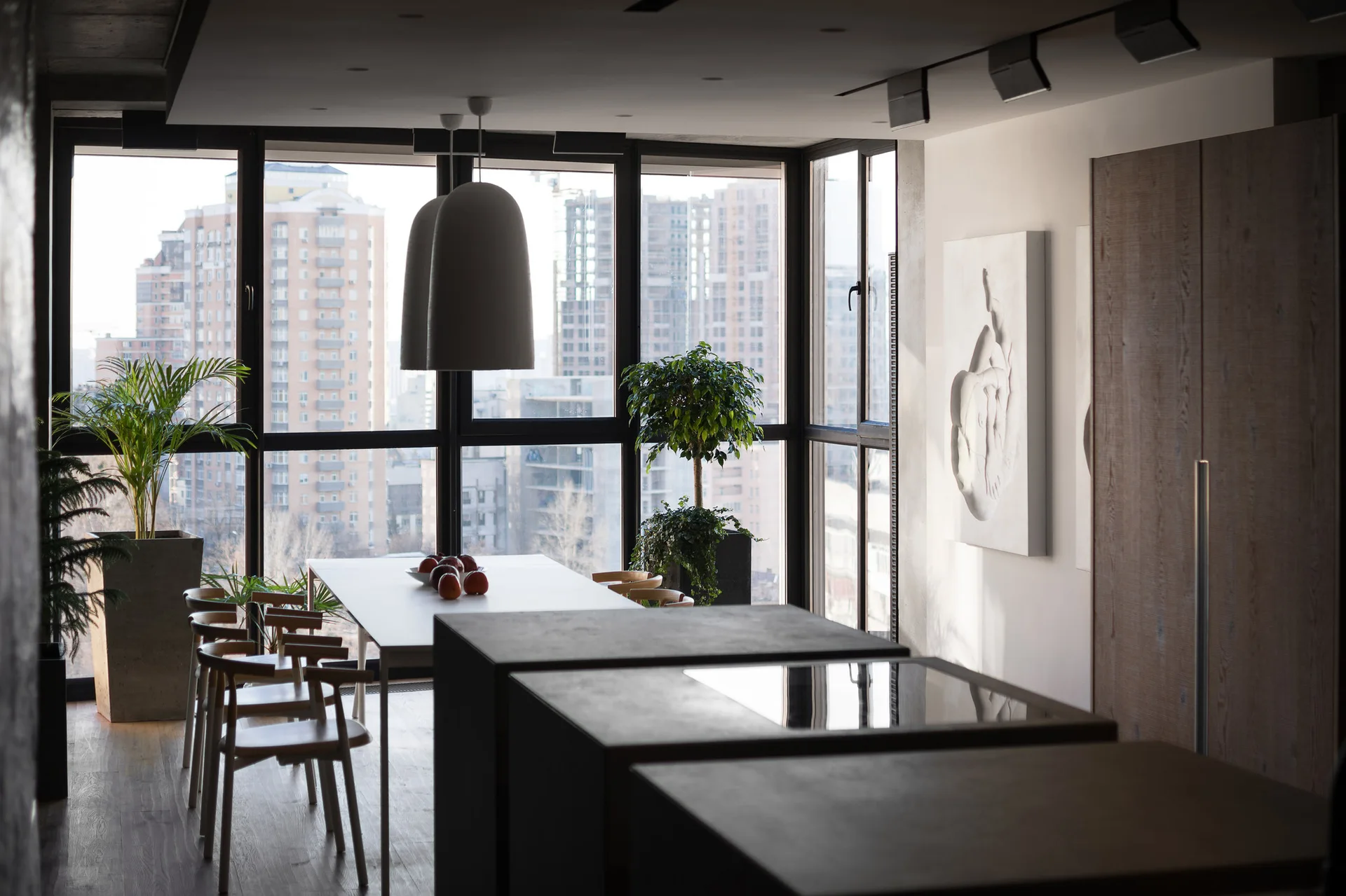 A modern dining room featuring a white table, wood chairs, a dark kitchen island, and a floor-to-ceiling window overlooking a city skyline.