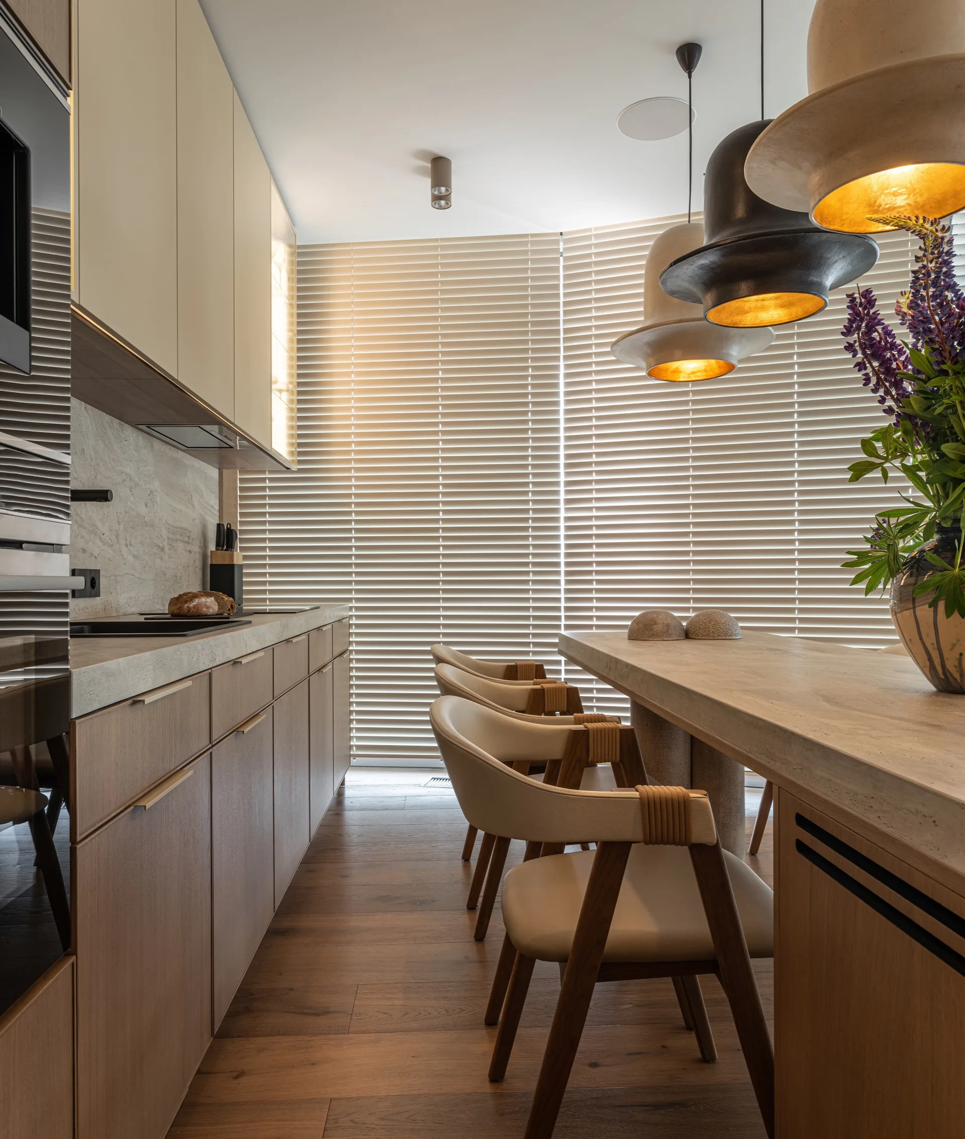 A modern kitchen features wood cabinets, a stone island with contemporary dining chairs, horizontal window blinds, and hanging pendant lights.