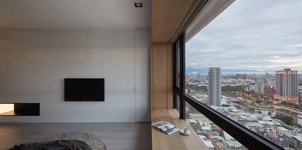A minimalist room features a wall-mounted television, gray flooring, and a large window with a built-in light wood window seat overlooking a city.
