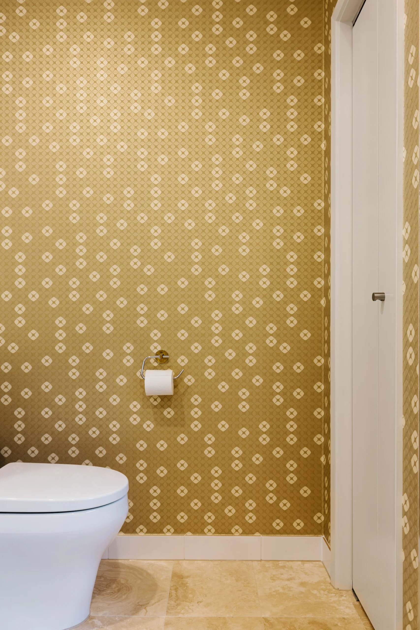 A small powder room featuring gold geometric patterned wallpaper, a white ceramic toilet, and light beige stone floor tiles.
