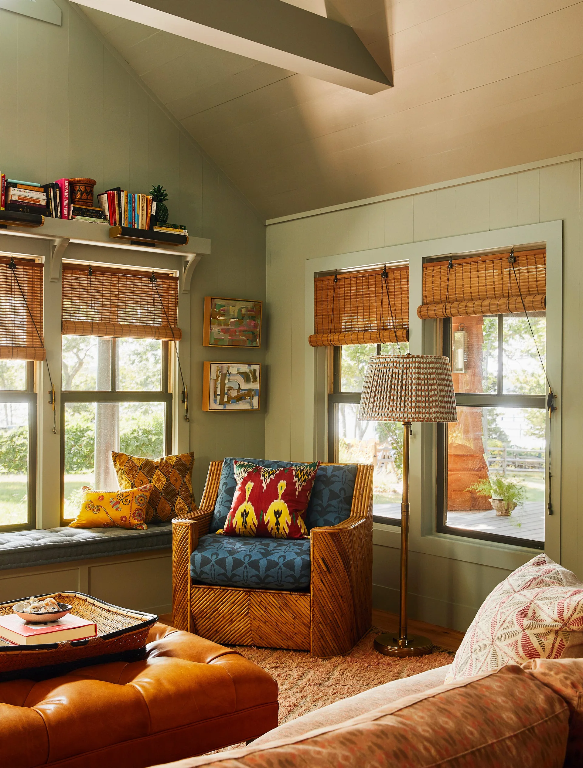 A cozy sage green family room featuring a patterned wicker armchair, a tufted leather ottoman, and windows with woven bamboo shades.