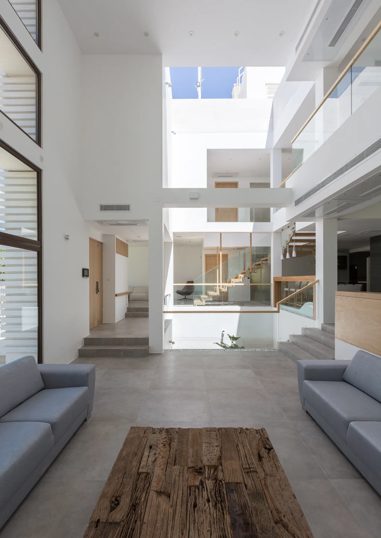 A modern open-plan living room with two gray sofas, a rustic wood coffee table, and a soaring white atrium with glass railings and stairs.