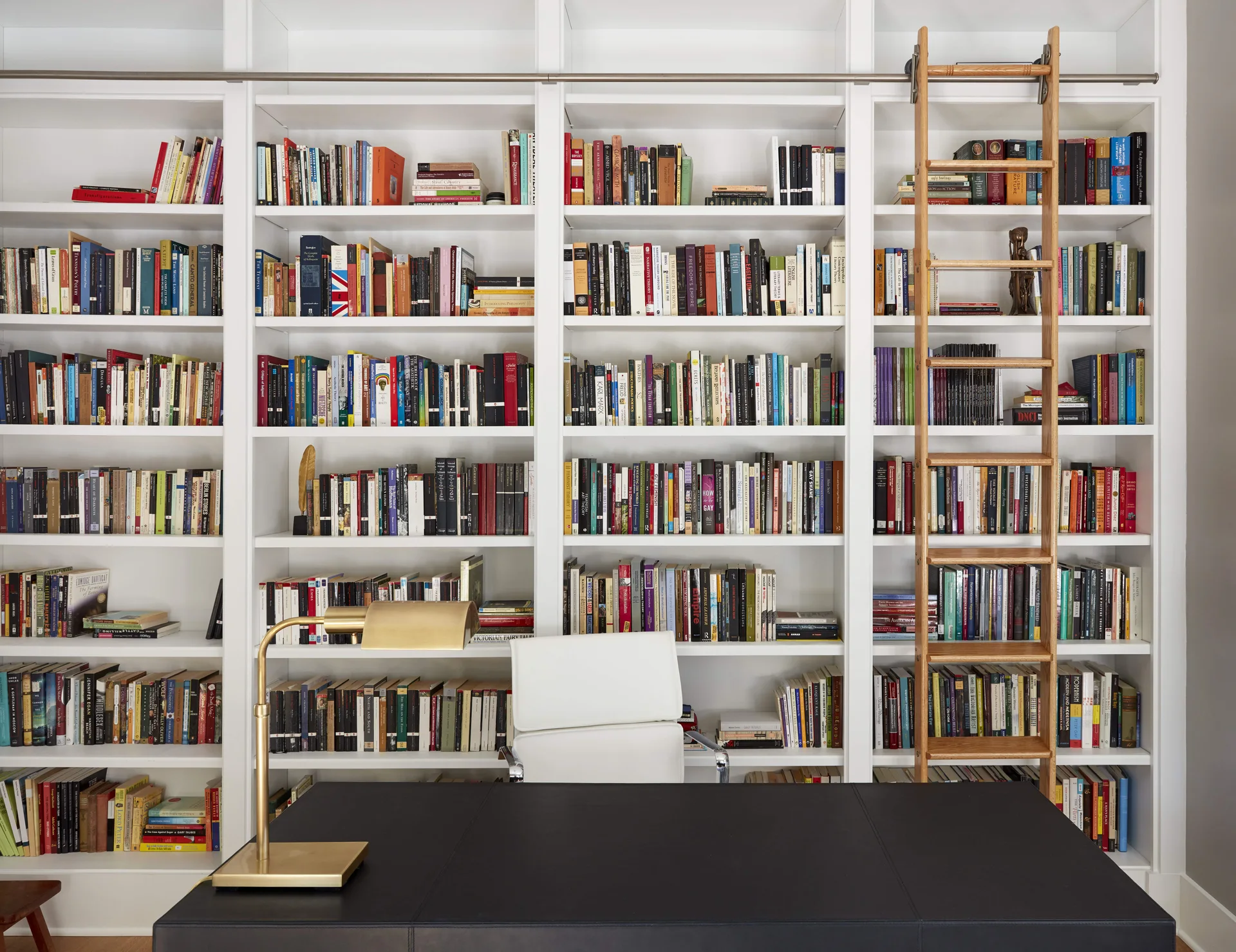 A home office library features a wall of white built-in bookshelves, a rolling wooden ladder, and a dark desk with a brass lamp.