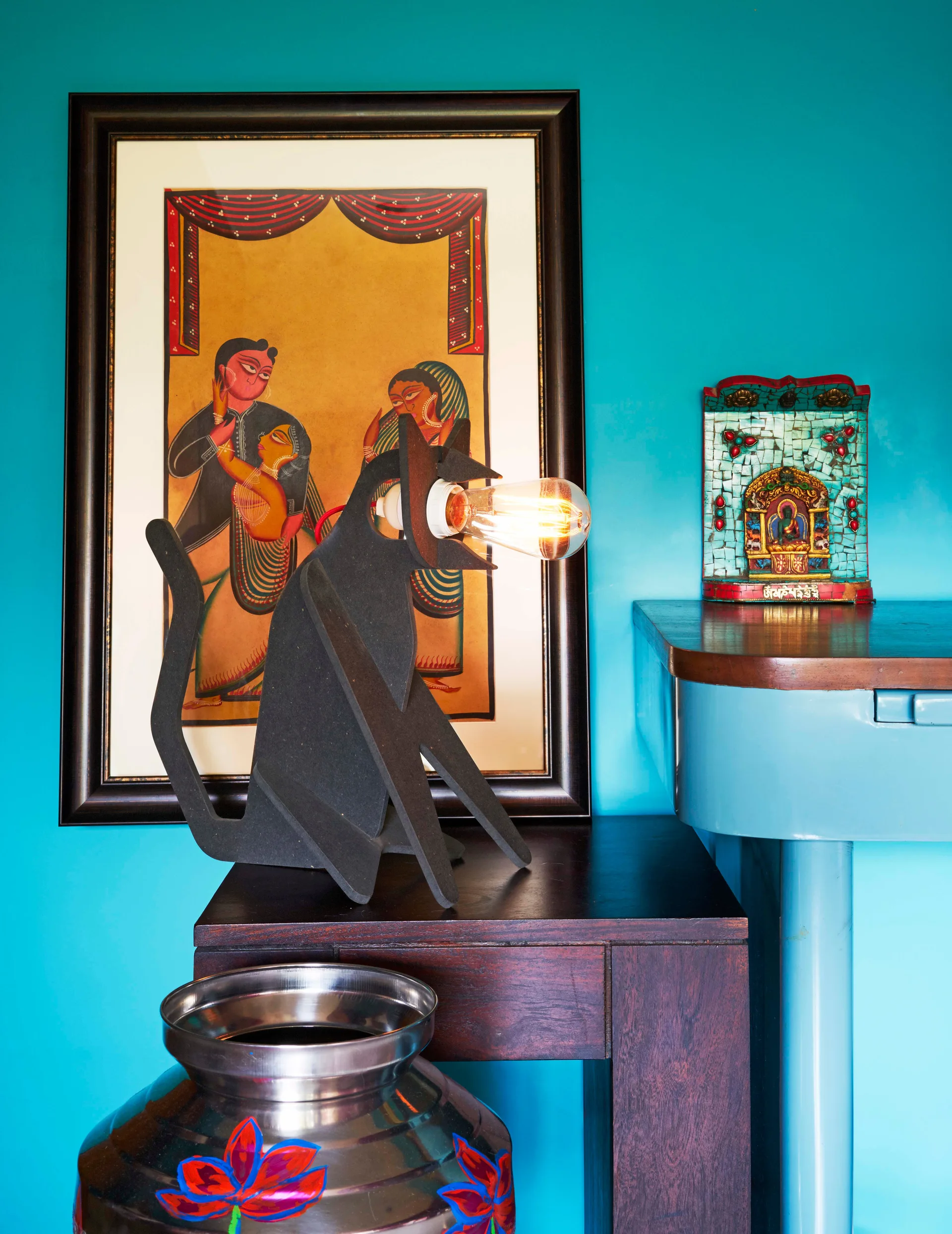 A black cat-shaped lamp with an exposed bulb sits on a dark wood table against a turquoise wall next to framed traditional art.