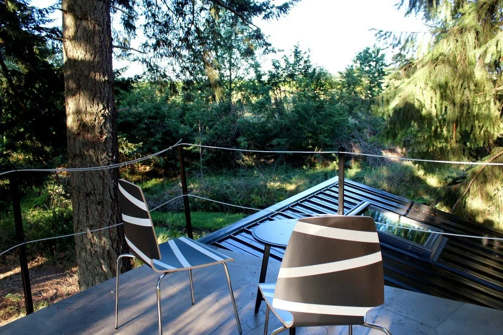 Two modern black and white striped chairs and a small table sit on a grey deck with a wire railing overlooking a dense green forest.