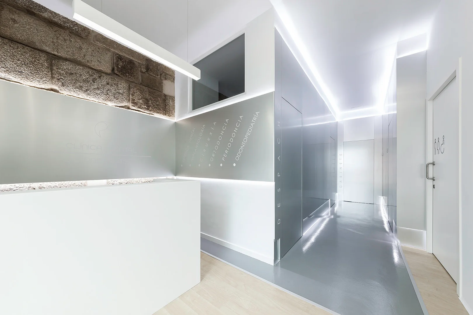 A minimalist commercial hallway features a white reception desk, reflective grey flooring, integrated linear cove lighting, and a patch of exposed stone wall.