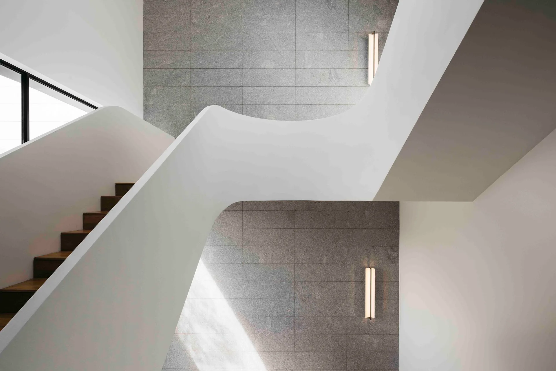 A sculptural white curved staircase with wood treads stands in front of a grey stone-tiled wall with two vertical light sconces.
