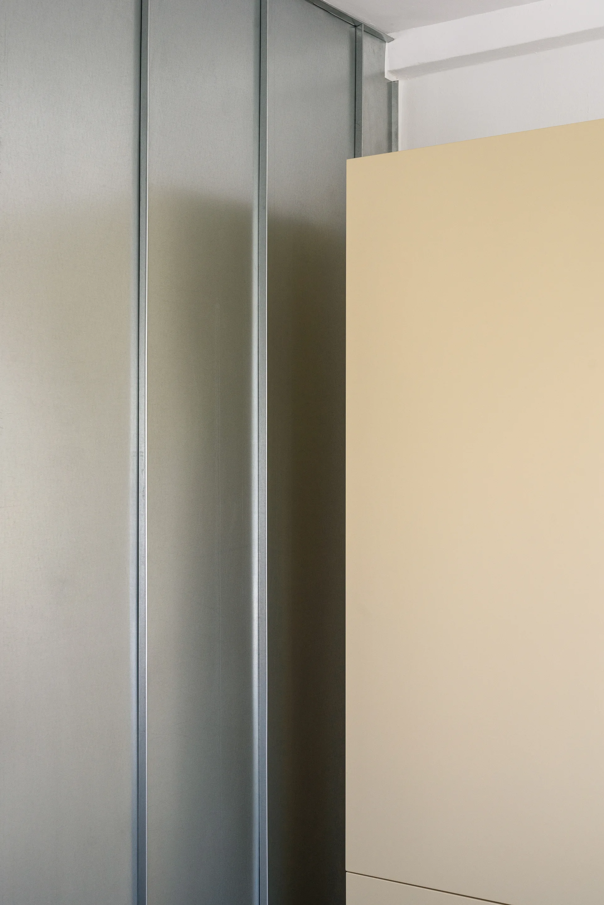 A close-up view of an interior wall junction showing cool grey metal panels with vertical seams next to a smooth, matte beige surface.