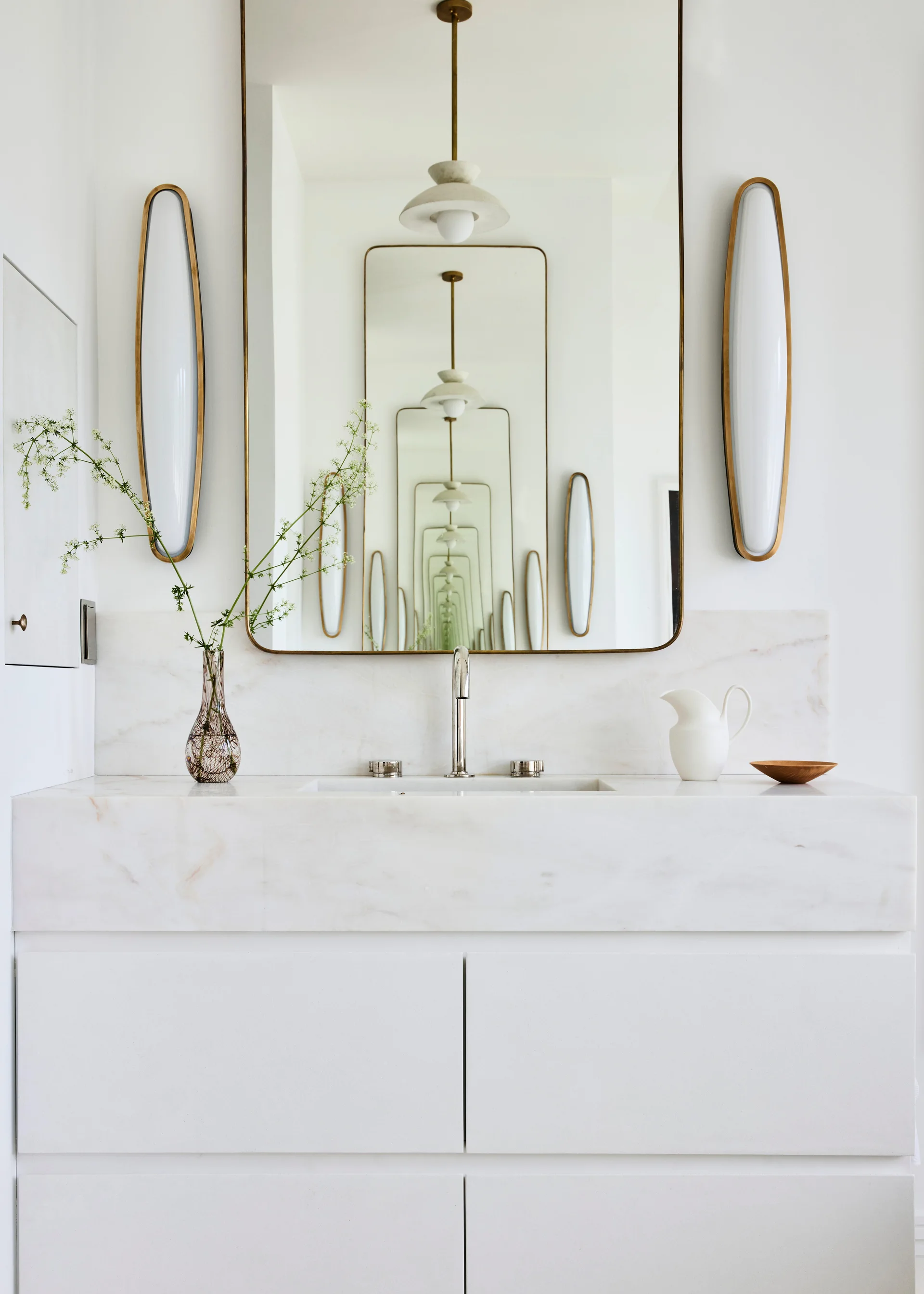 A white bathroom vanity features a marble countertop, silver faucet, a rectangular brass-framed mirror showing infinite reflections, and two capsule sconces.