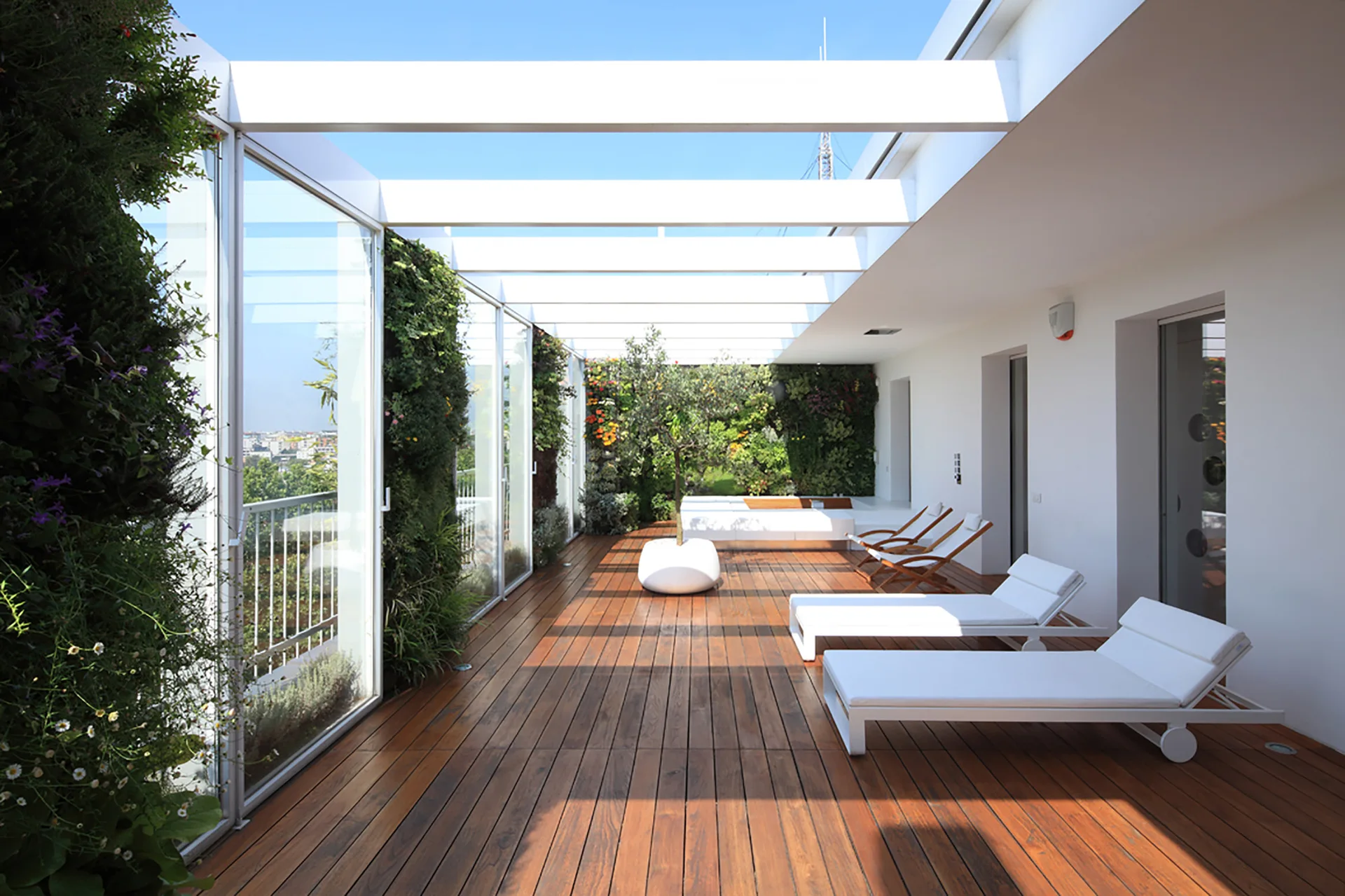 A long wooden deck terrace features white minimalist lounge chairs, a white structural pergola overhead, and green vertical gardens lining one side.