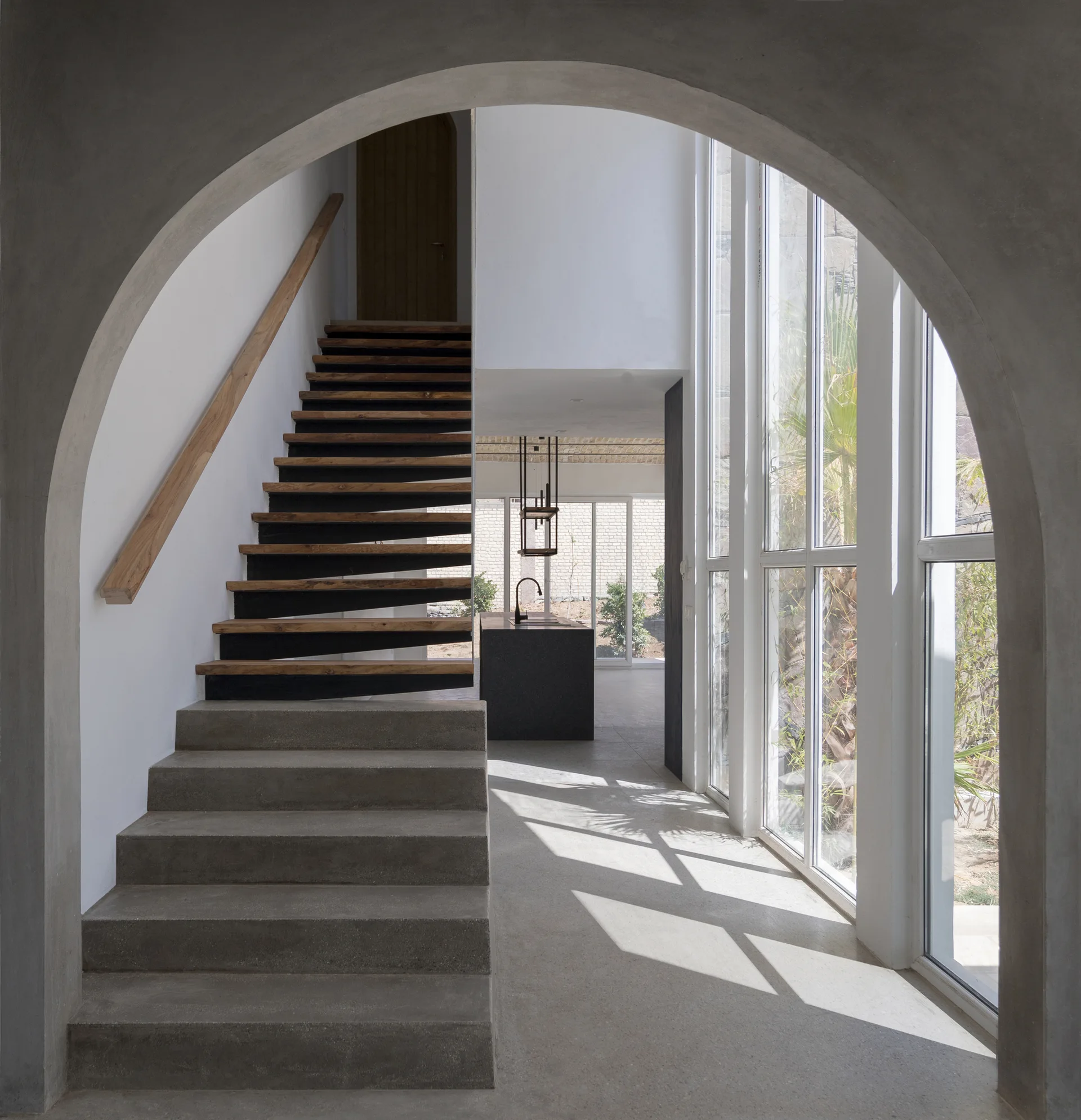 A minimalist home hallway viewed through a concrete arch, showing a floating wooden staircase and tall windows.