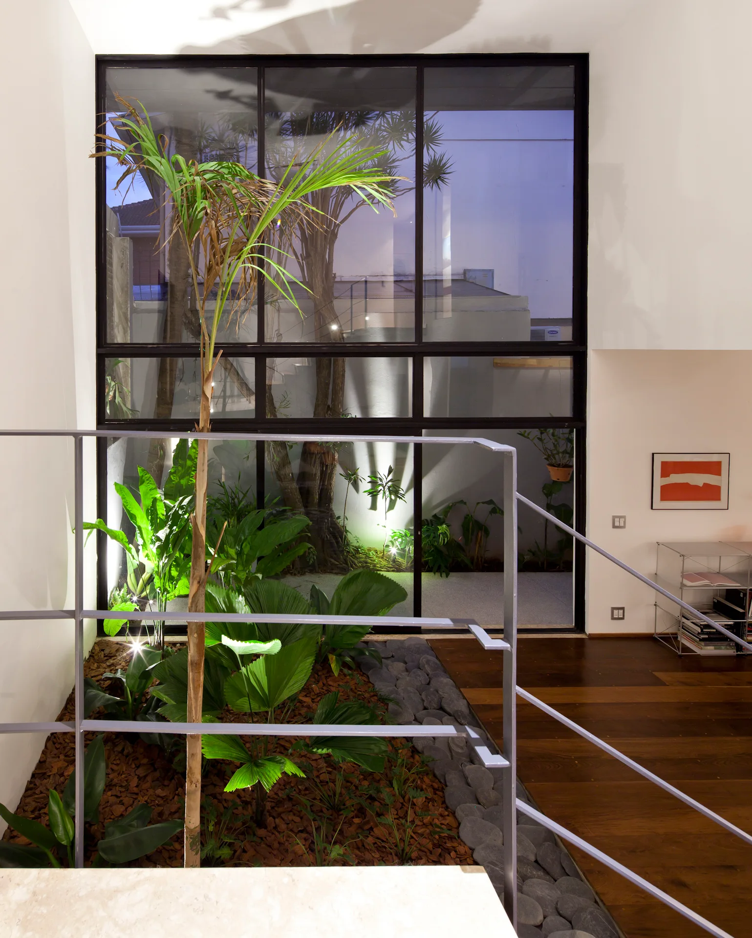 A modern home interior features an indoor atrium garden with tropical plants and river stones, visible through a black metal framed glass wall.