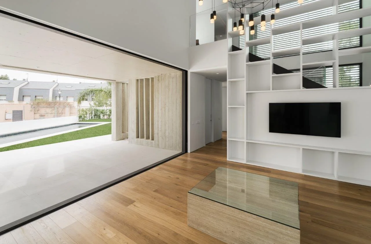 A minimalist living room with wooden flooring features a large white built-in shelving unit, a stone coffee table, and sliding glass doors to a patio.