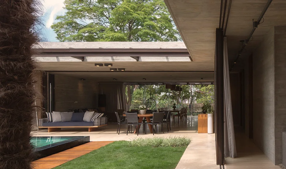 An open-air concrete pavilion features a lounge area with a navy sofa, a wooden dining table, and an adjacent lawn and swimming pool.